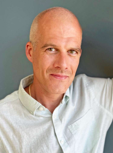 A man with a shaved head and blue eyes, wearing a light-colored button-up shirt, smiling slightly, with a gray background.