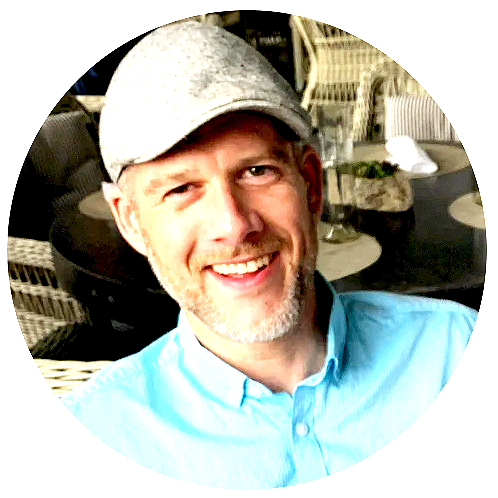 A man with a beard and light-colored hair wearing a light blue shirt and a gray cap, smiling and winking, seated outdoors with furniture and plants in the background.