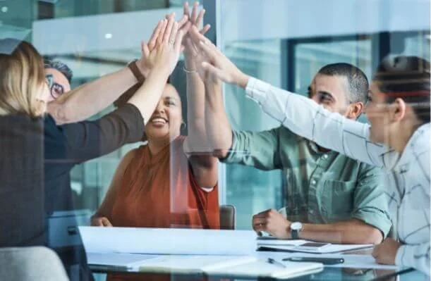 A group of diverse colleagues in a meeting giving each other high fives in an office setting.