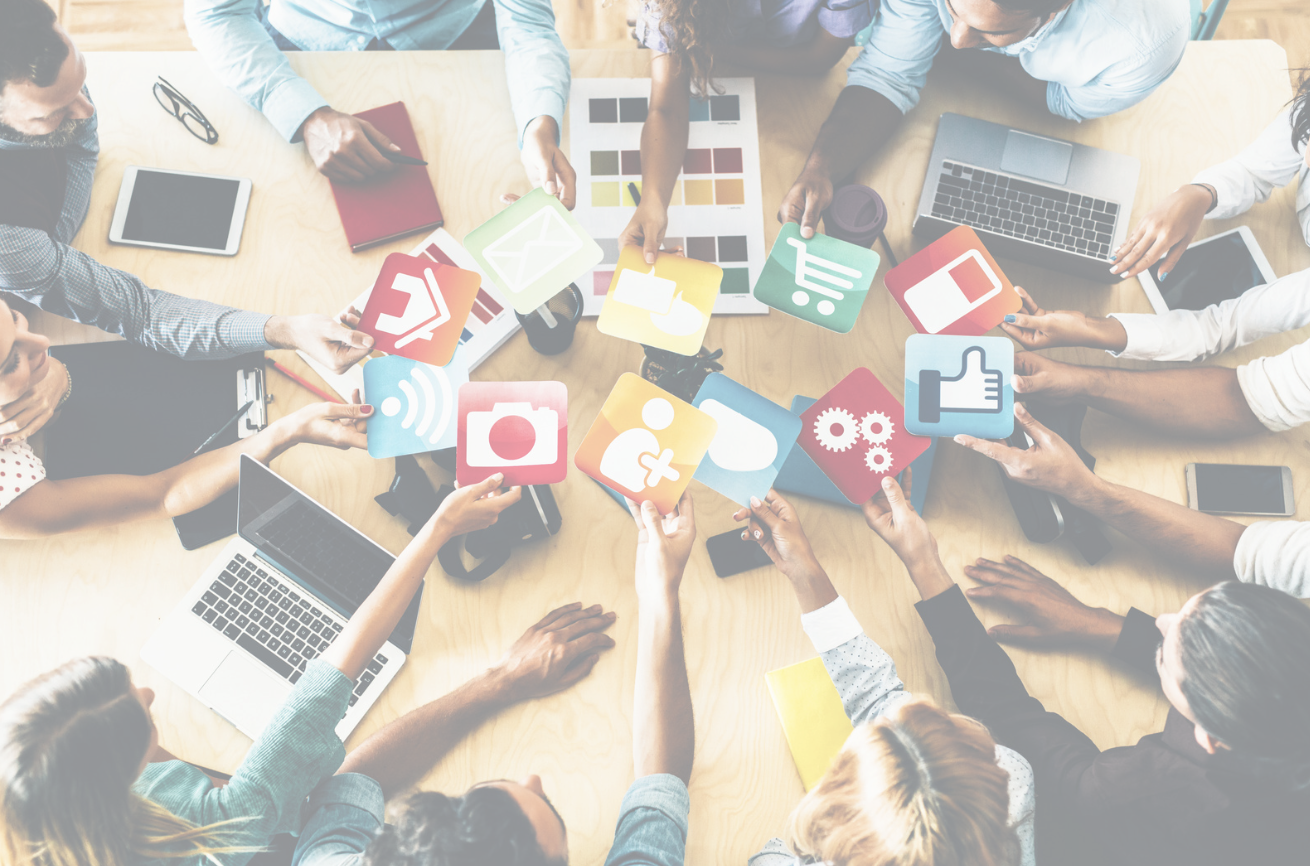 Group of people sitting around a table, holding colorful icons representing social media and digital content, with laptops and tablets on the table.