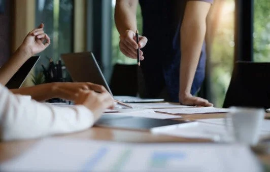 Businesspeople in a meeting, with one person standing and writing, another person sitting and working on a laptop, and documents on the table.