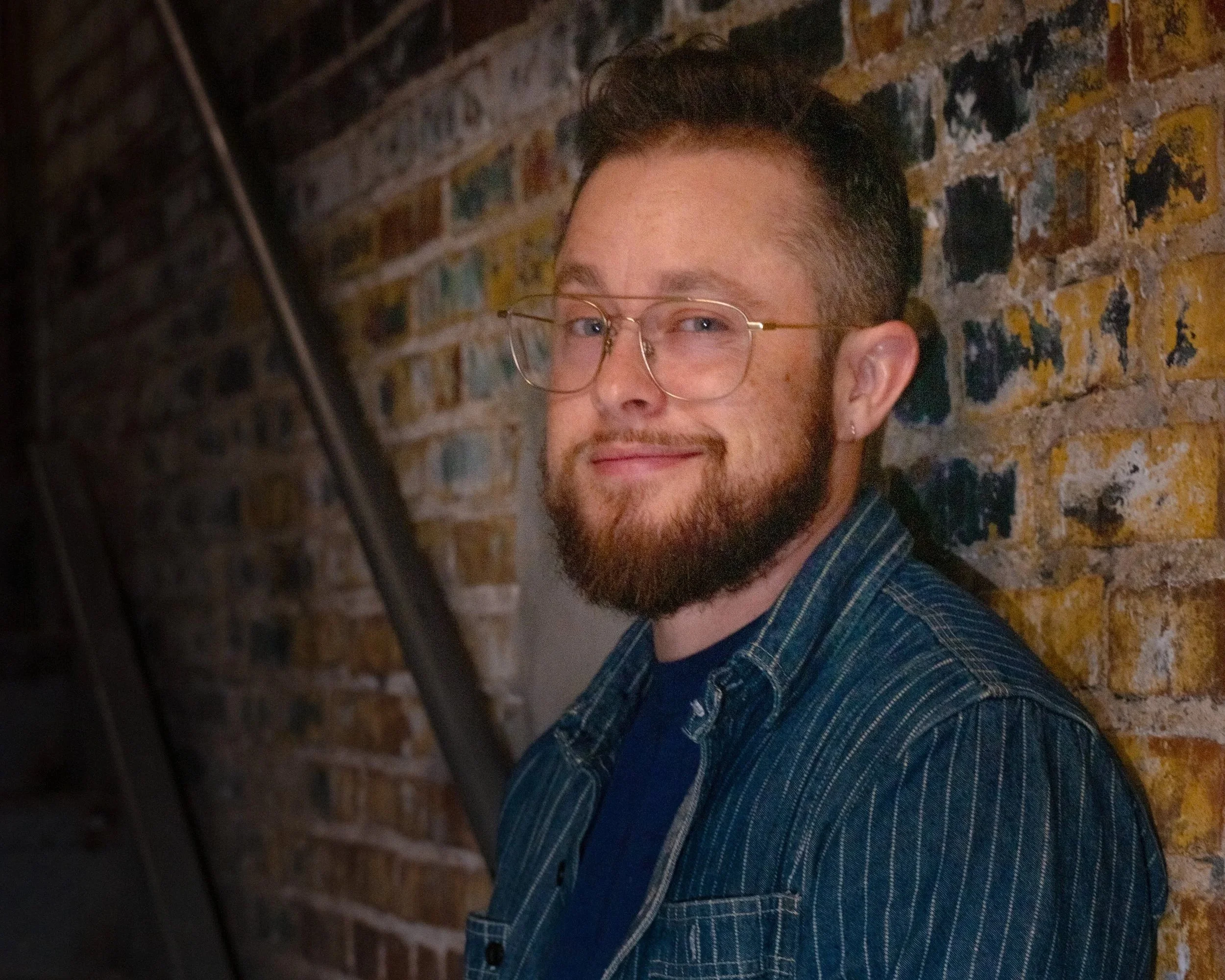River Butcher leaning against a brick wall, wearing a denim jacket over a dark shirt, smiling softly.