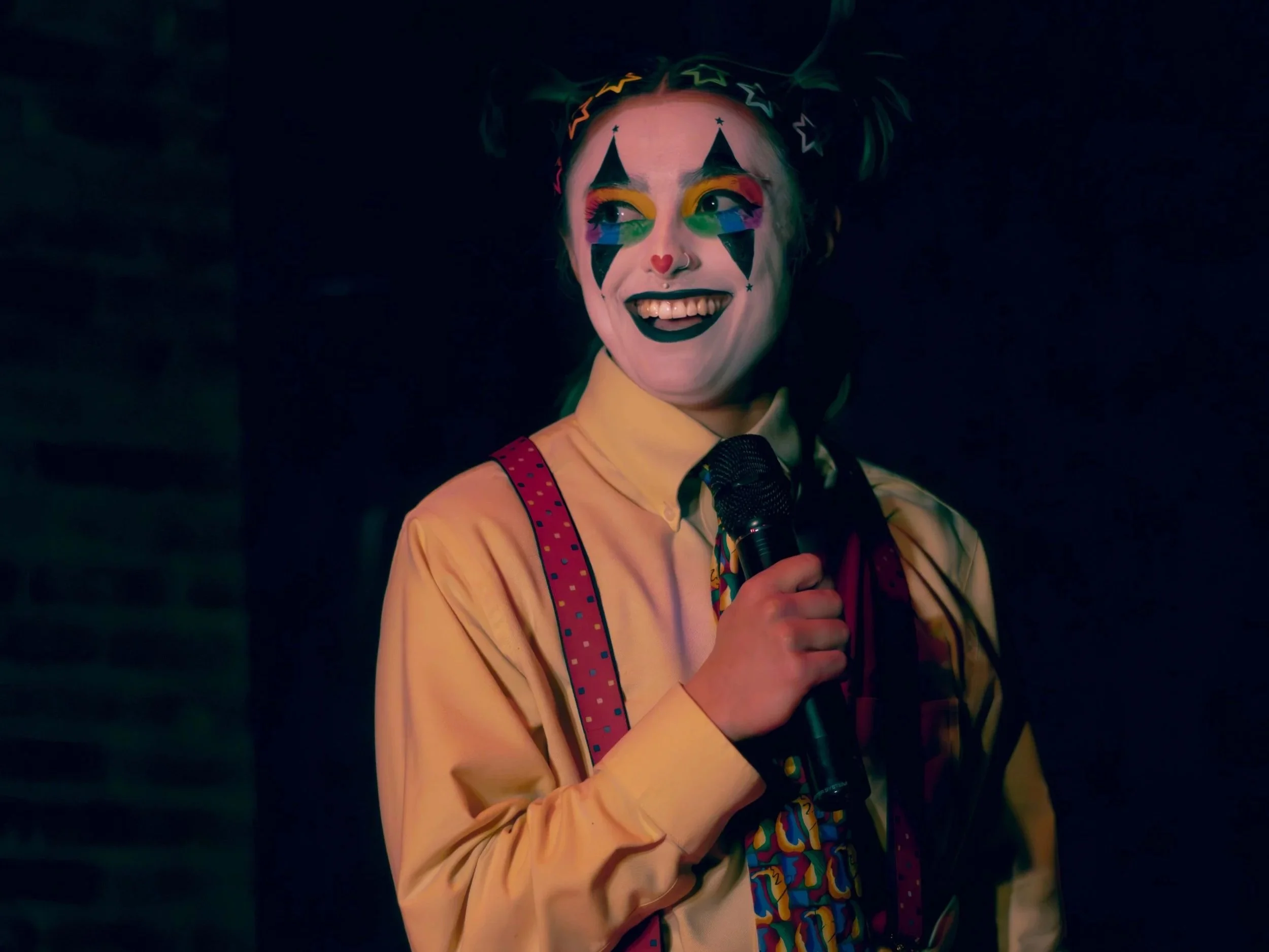 Clown with colorful makeup, a yellow shirt, and holding a microphone, smiling in a dark setting.