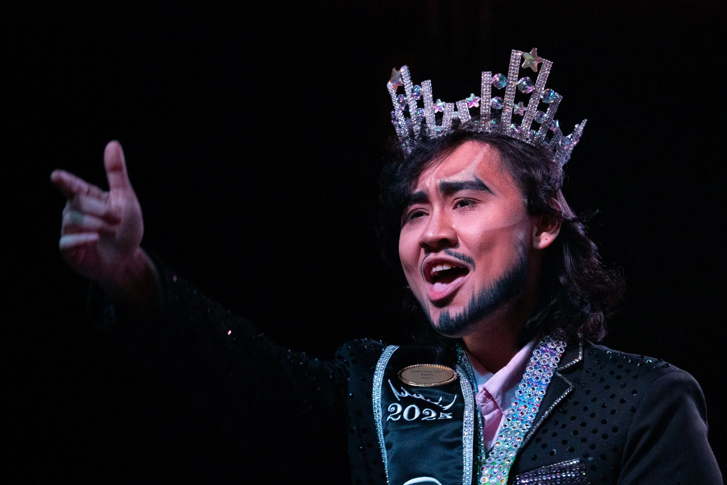 A performer with a shiny crown, makeup, and a costume on stage with a dark background.