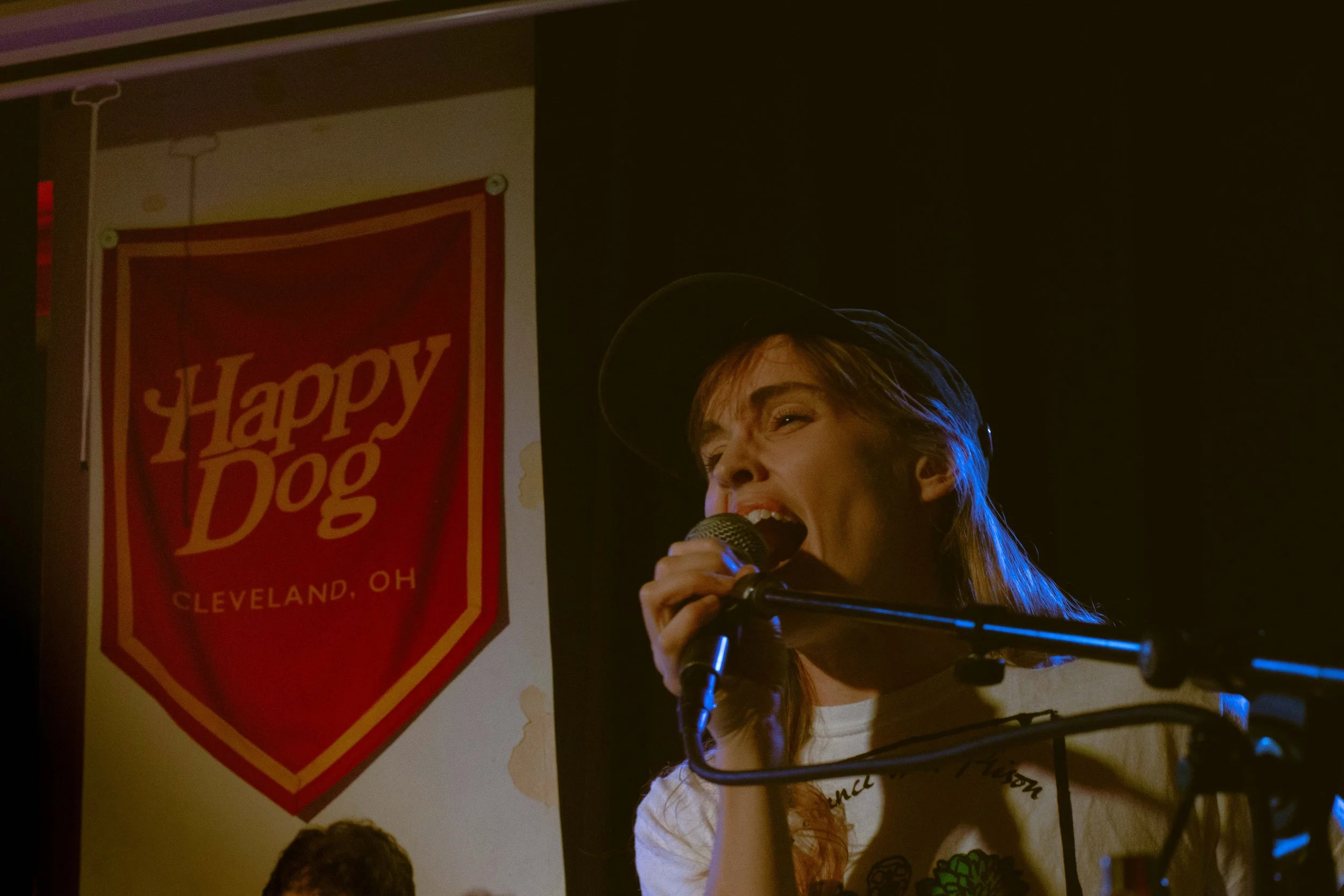 Singer with blonde hair, wearing a baseball cap, singing into a microphone at a concert. There is a red flag/banner with the words 'Happy Dog' and 'Cleveland, OH' visible in the background.