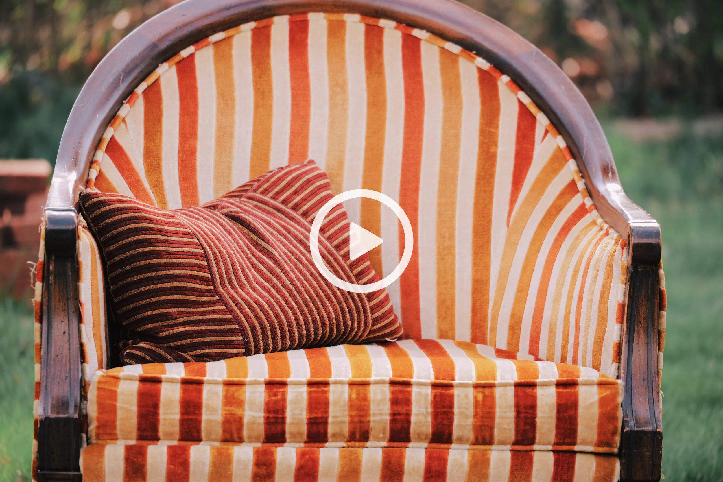 A vintage outdoor porch chair with a striped orange and beige cushion and a matching striped pillow, set against a blurred natural background.