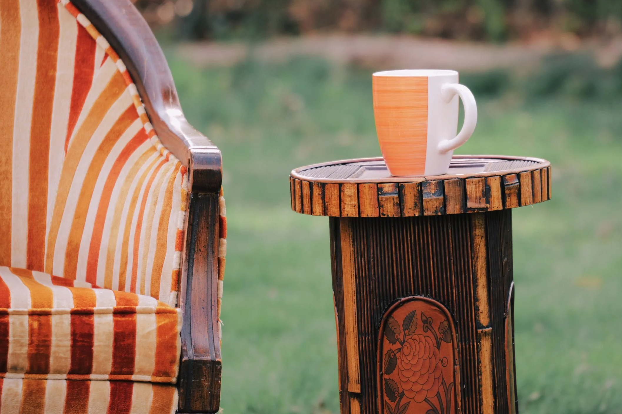 A colorful outdoor chair with orange and yellow striped upholstery next to a tall wooden side table holding a large ceramic mug with an orange stripe. In the background, there is a blurred outdoor landscape with grass and trees.