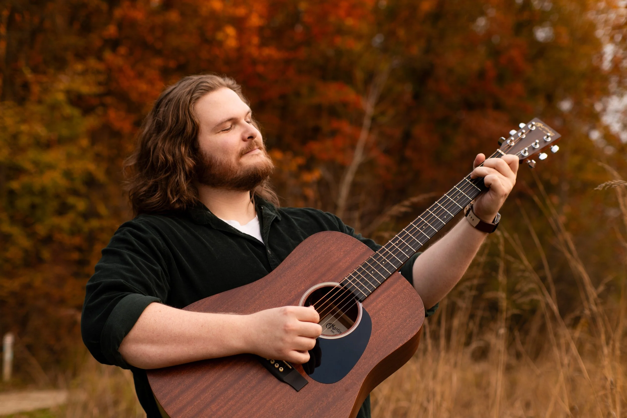 A man with long hair and a beard playing an acoustic guitar outdoors during autumn, with red and orange foliage in the background.