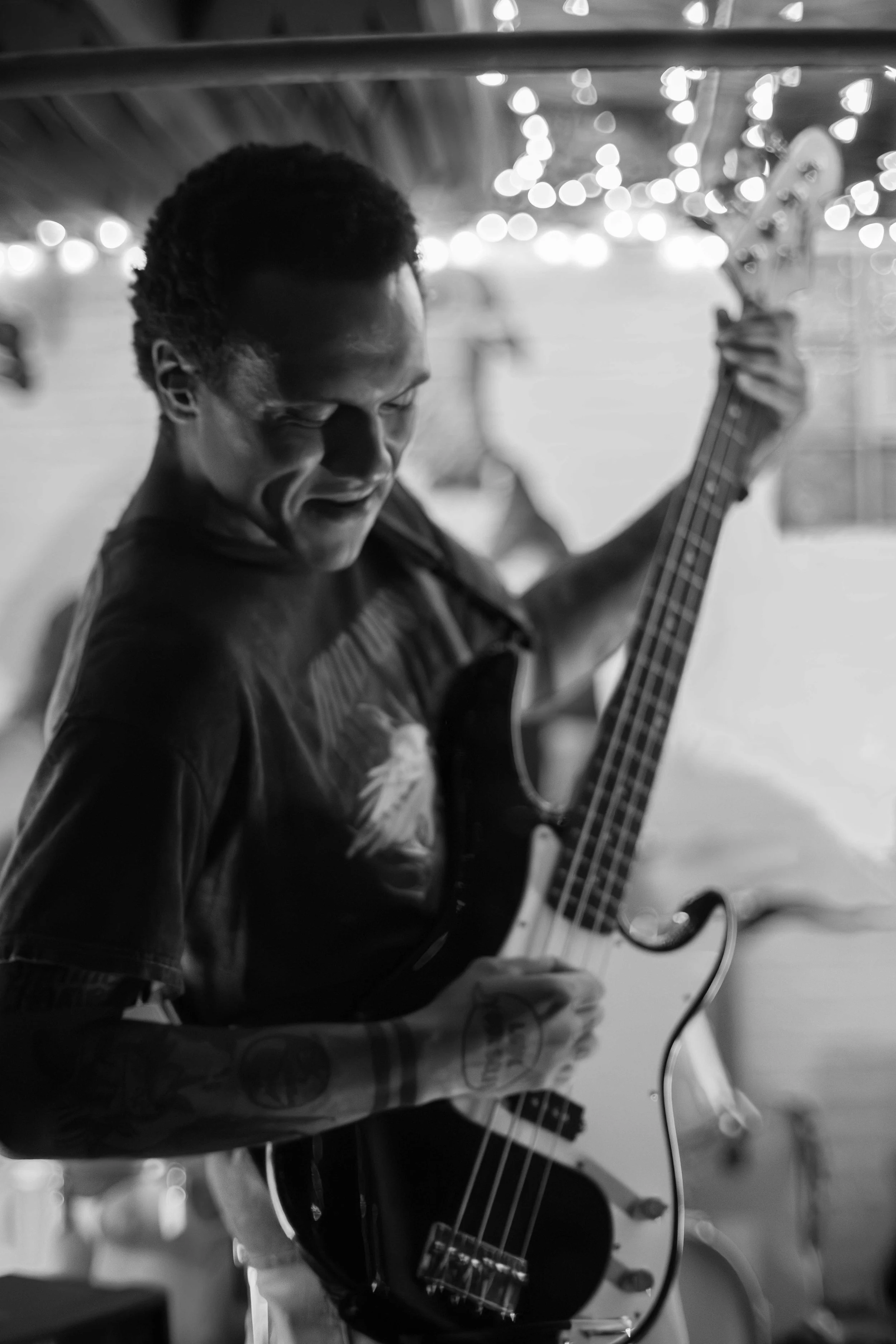 A person with short curly hair playing an electric guitar, with bright string lights in the background, in a black-and-white photograph.