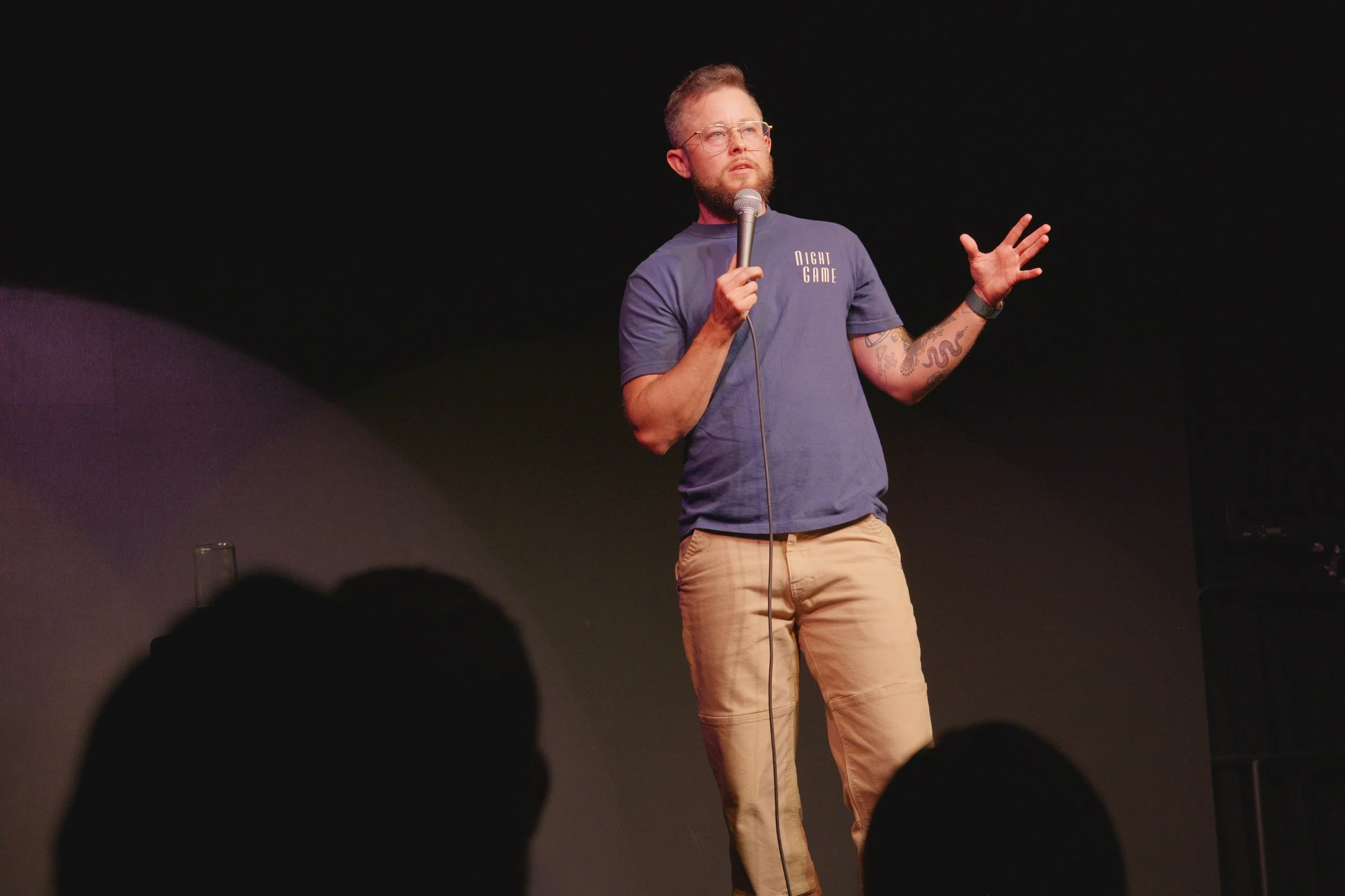 A man with glasses, a beard, and tattoos on his left arm is holding a microphone and speaking on stage. He is wearing a blue T-shirt with the words 'Night Game' printed on it and tan pants, with a black wristband on his right wrist. The stage has a d