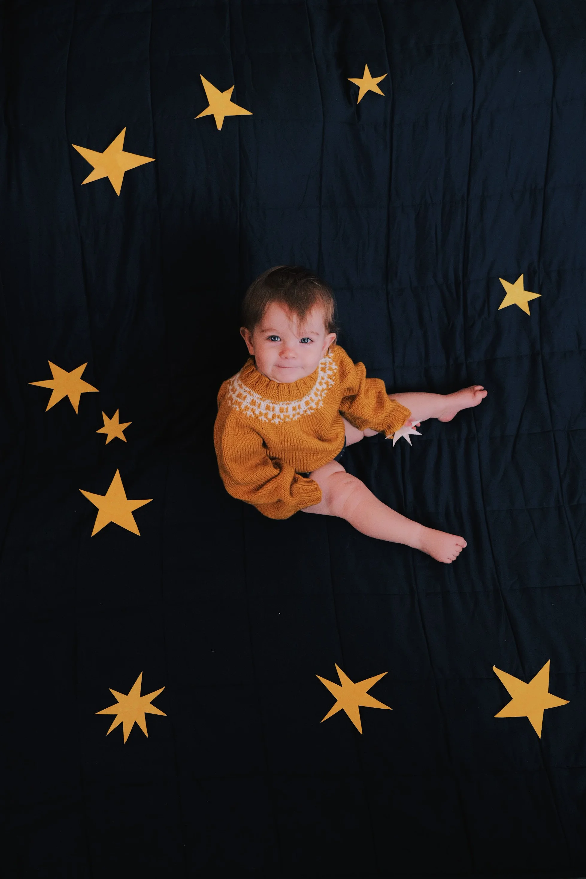 A young child with brown hair and blue eyes is sitting on a black quilted fabric with yellow paper stars attached. The child is wearing a mustard colored sweater with a white pattern across the chest.
