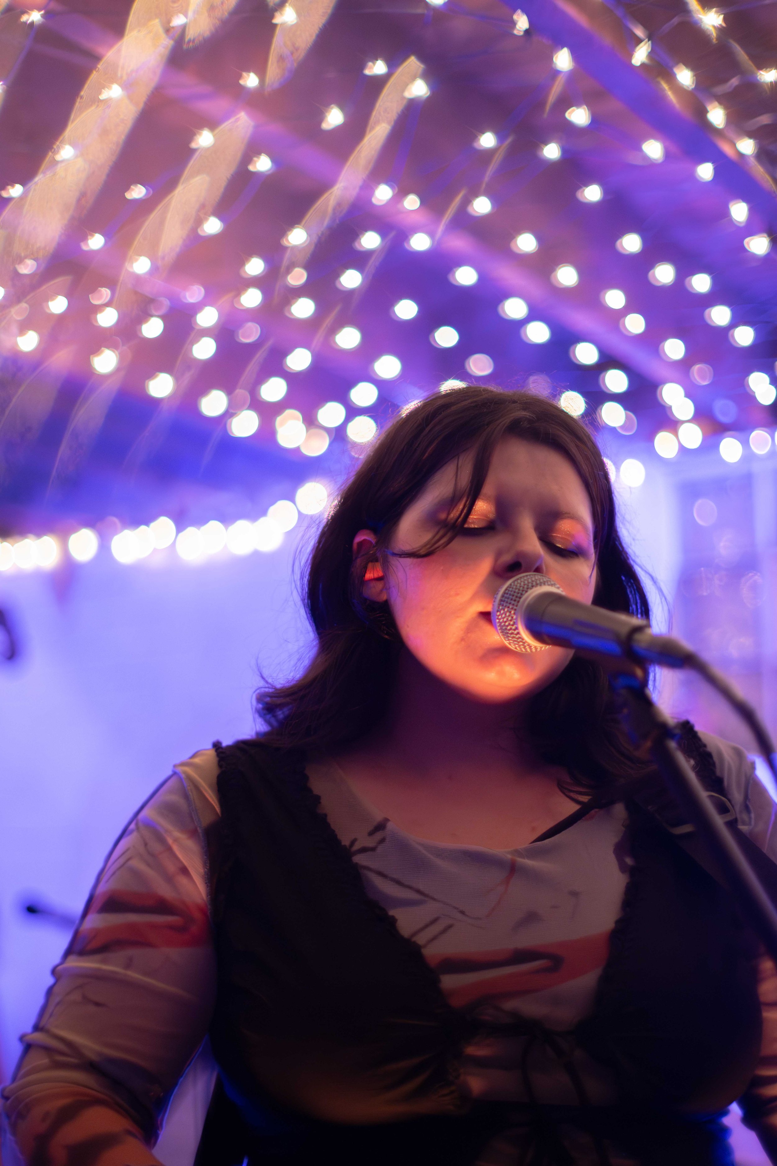 Singer singing into a microphone with closed eyes in a dimly lit room with purple and pink string lights overhead.