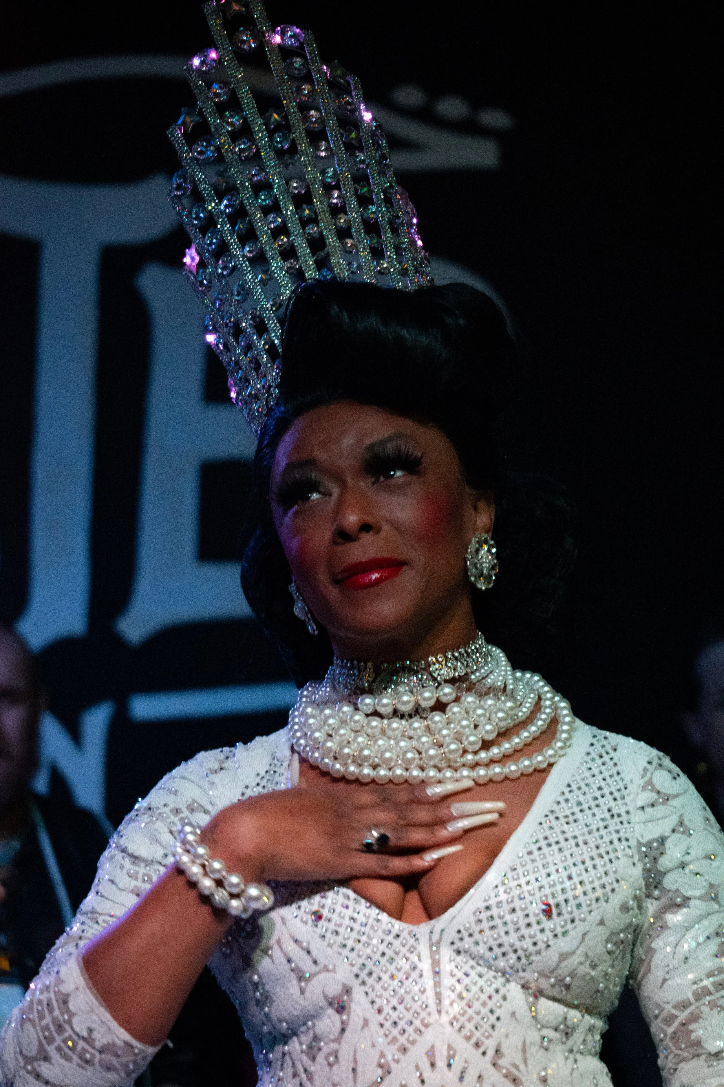 A woman dressed in elegant white attire with multiple pearl necklaces, large pearl earrings, and a floral bracelet, standing against a dark background. She is wearing a large, sparkling tiara and has her hand on her chest.