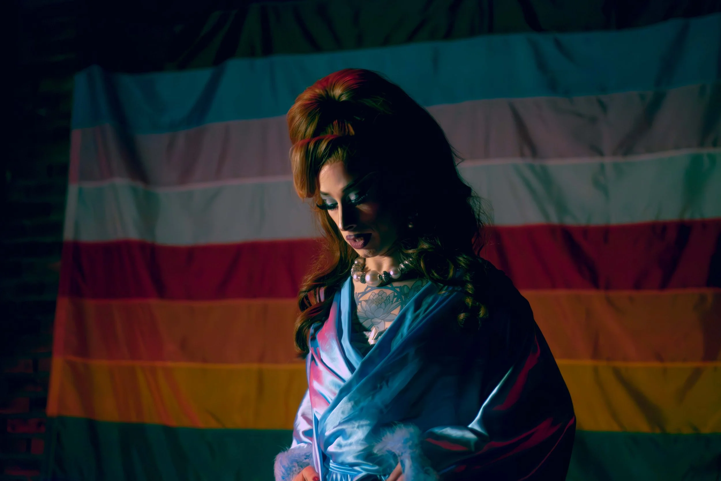 Drag queen with long hair and makeup looking down, standing in front of a flag with horizontal stripes of blue, white, and red, illuminated by colorful lights.