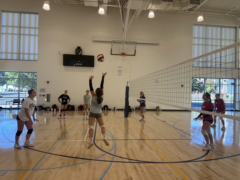 A women's volleyball game in progress in an indoor gym. One player is jumping to hit the ball over the net while others are ready to receive or defend.