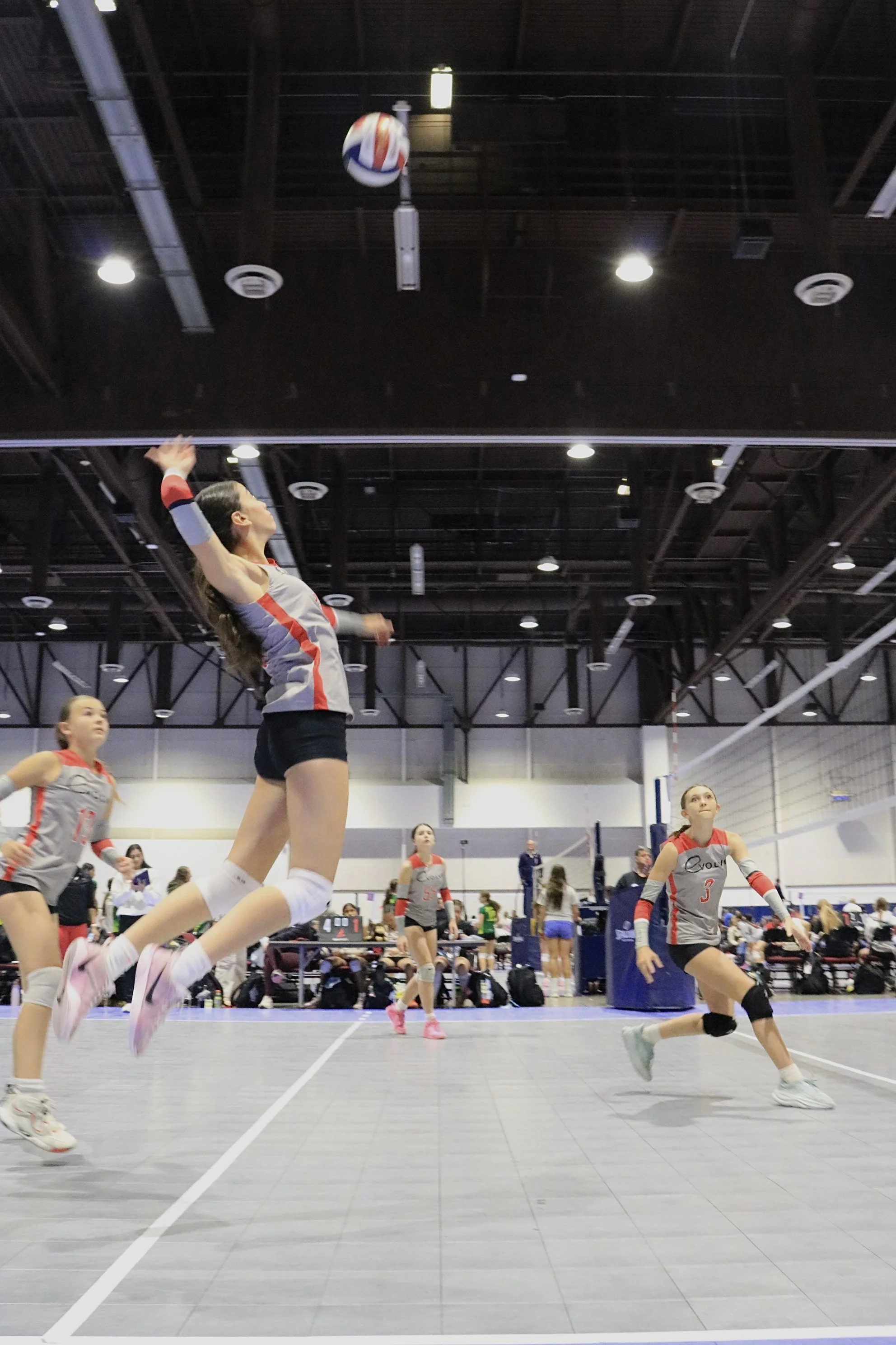 A volleyball game in progress at an indoor gymnasium with volleyball players jumping, running, and preparing to hit the ball, which is mid-air above the net. The players are dressed in sports uniforms, and the gym has a high ceiling with bright overhead lights.