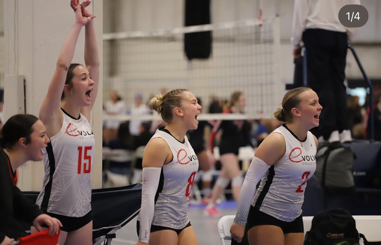 Four female volleyball players in team uniforms celebrating on the sidelines of a volleyball court during a match.