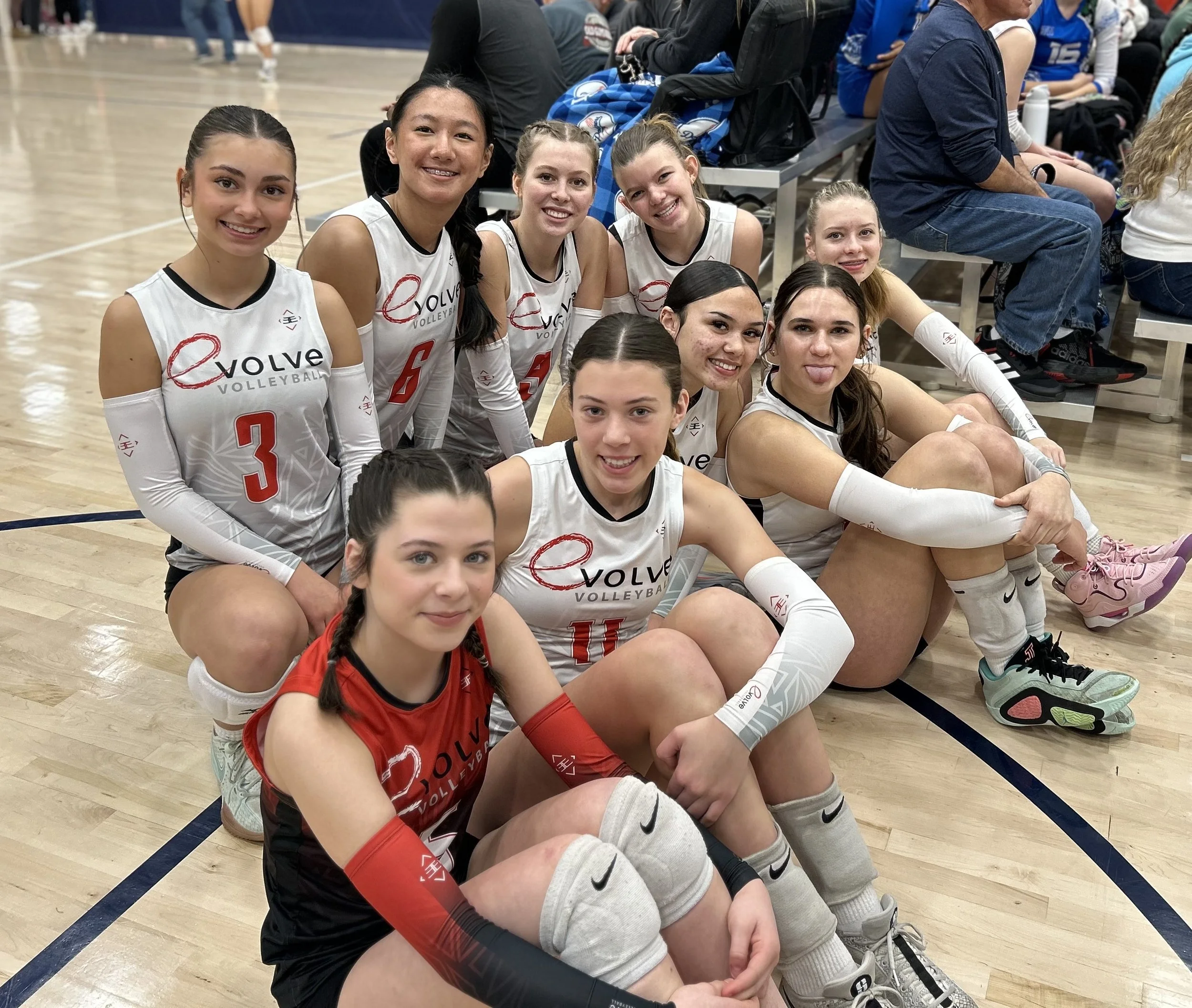A group of young female volleyball players sitting on the floor of an indoor gym, smiling and posing for a photo. They are wearing white volleyball uniforms with the logo 'Evolve Volleyball' and red numbers, and some are wearing knee pads and athletic shoes.
