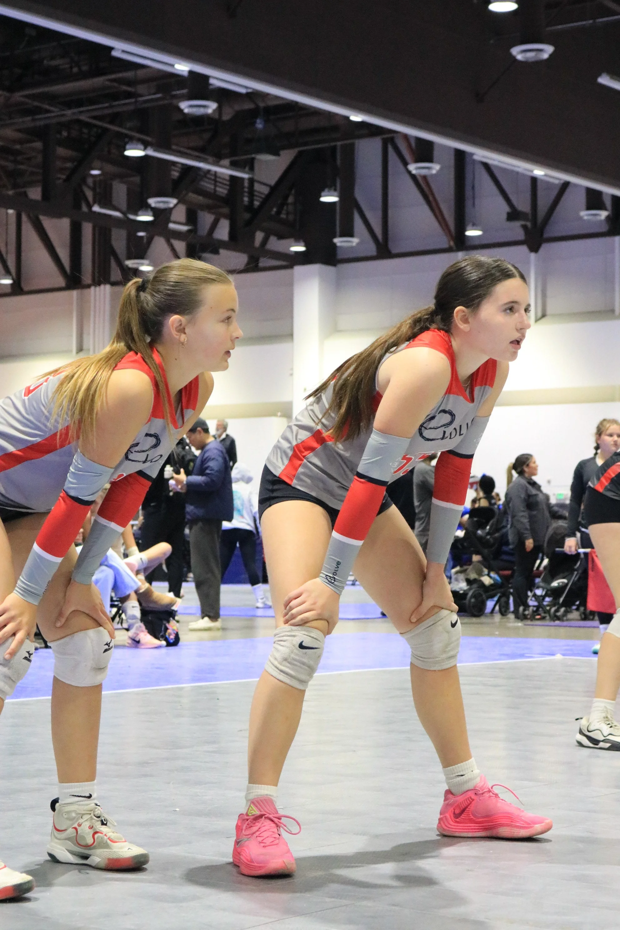Two female volleyball players in red and gray uniforms crouched on the court during a game, with a busy indoor sports arena and spectators in the background.