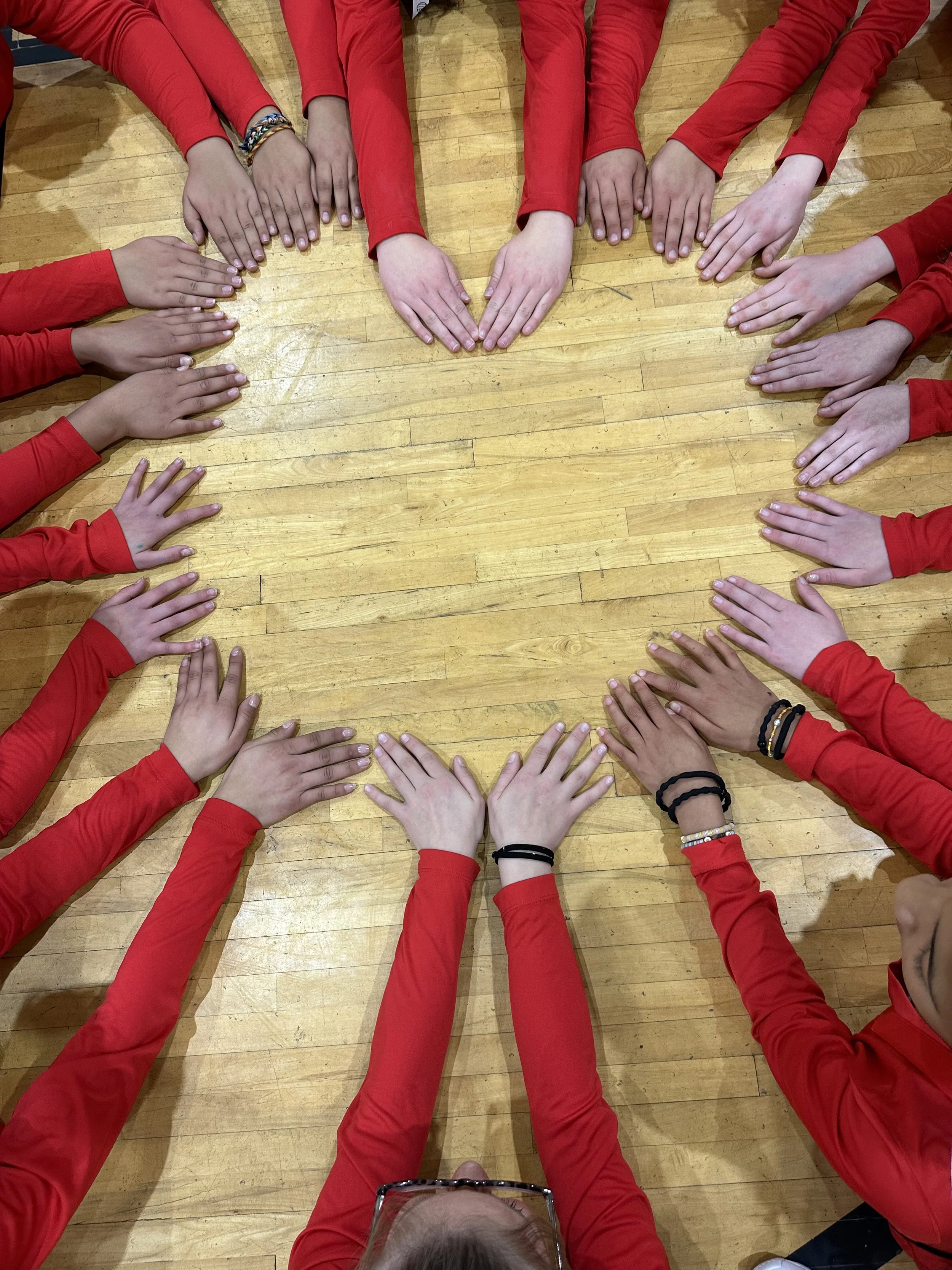 Group of people wearing red shirts sitting around a wooden table with their hands together, forming a heart shape in the center.