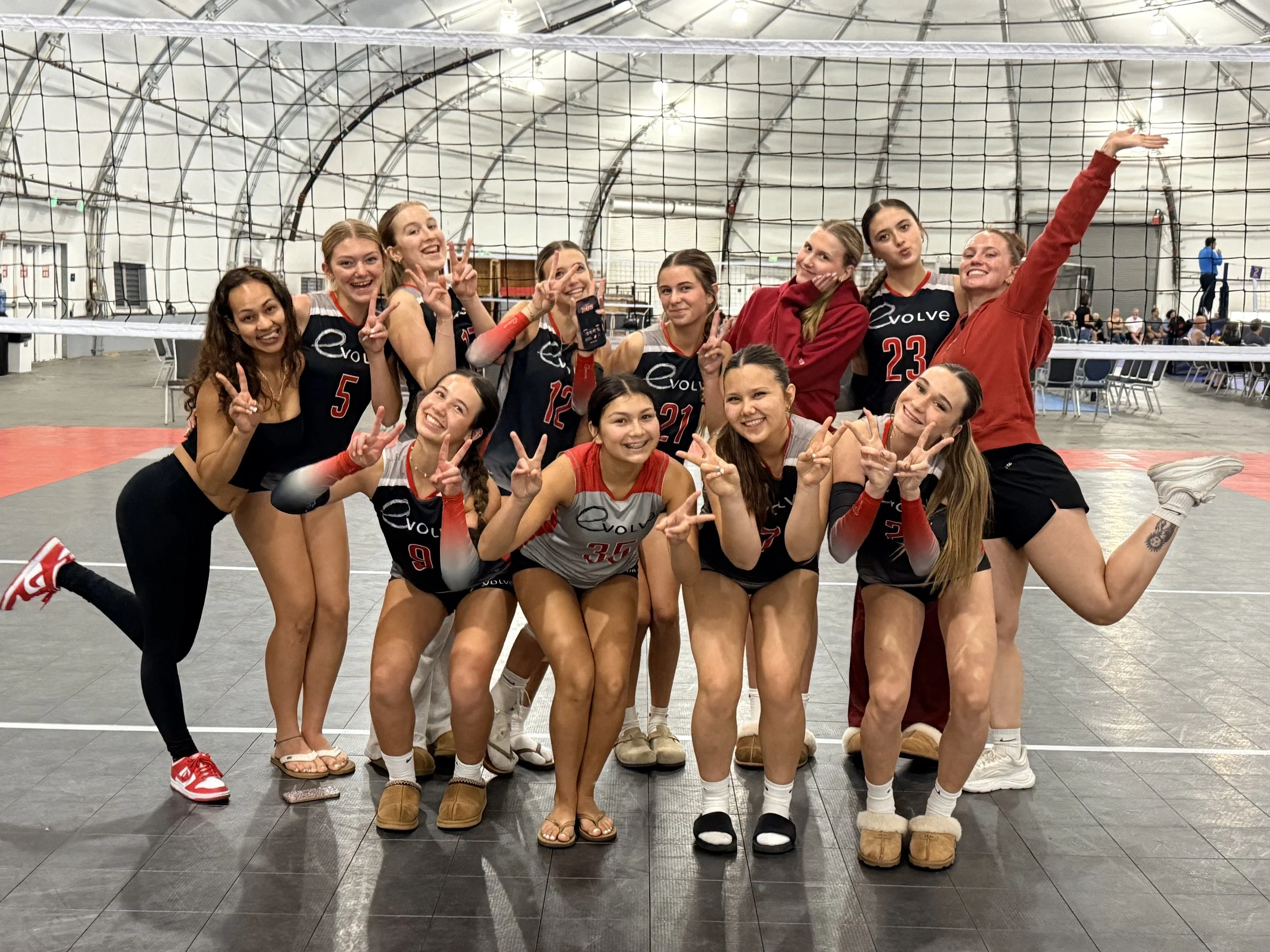 Group of young women in volleyball uniforms posing inside an indoor sports arena with a volleyball net behind them. They are smiling, making peace signs, and appear to have finished a game or practice.