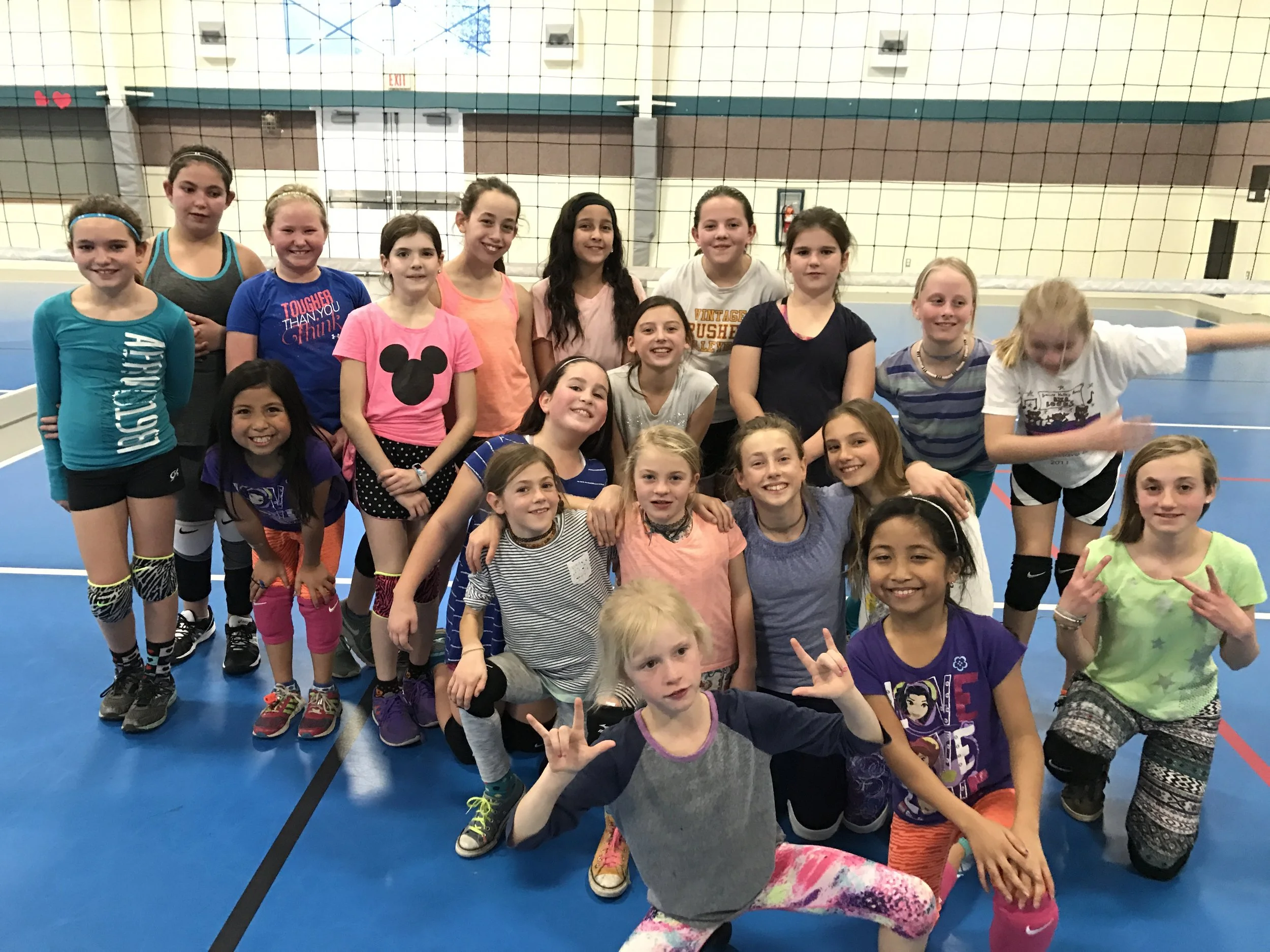 A group of young girls is gathered together in an indoor gymnasium, smiling and posing for a photo. They are wearing athletic clothing and standing in front of a volleyball net.