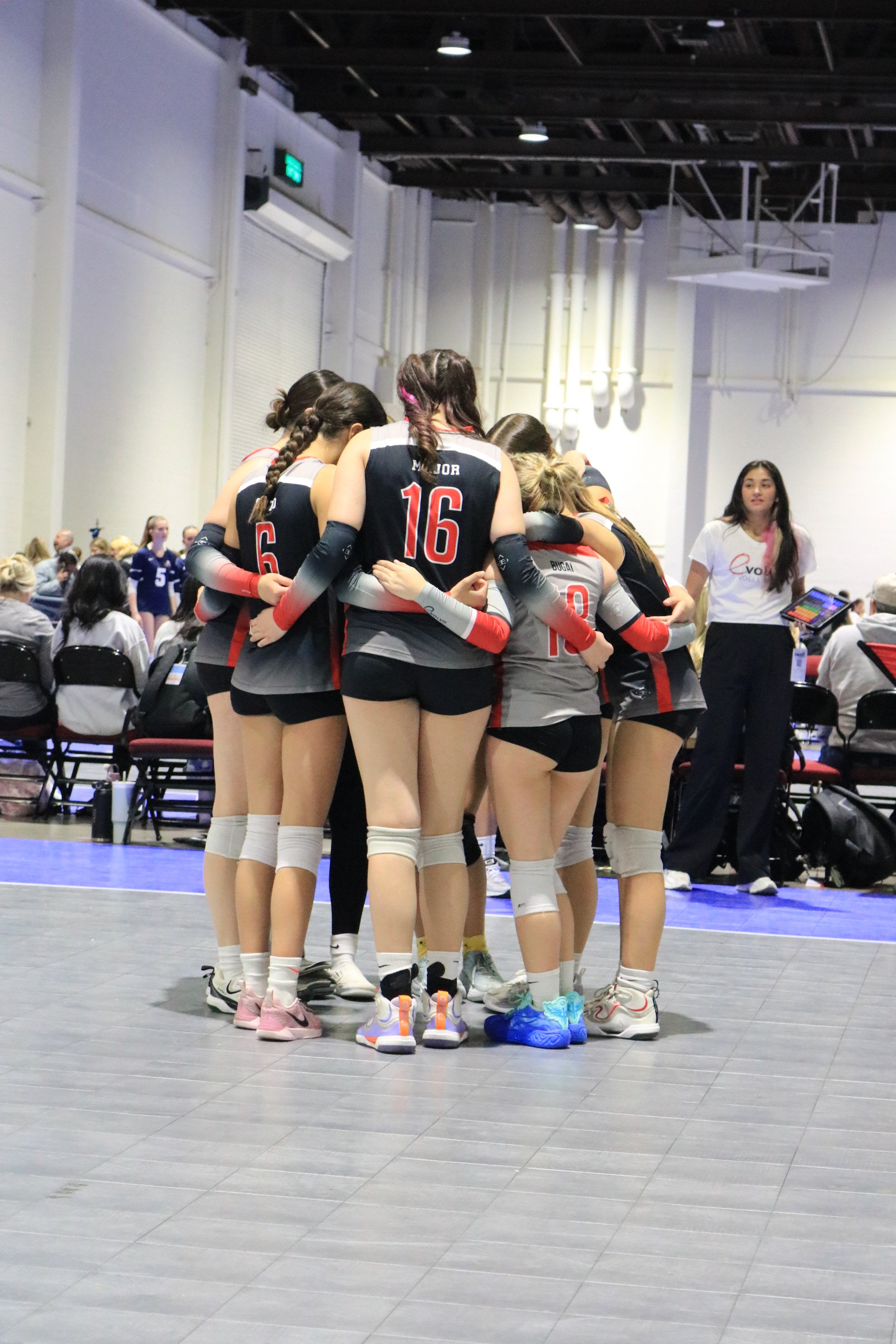 A volleyball team of young women huddles together on an indoor court during a game or practice, with a coach standing nearby.