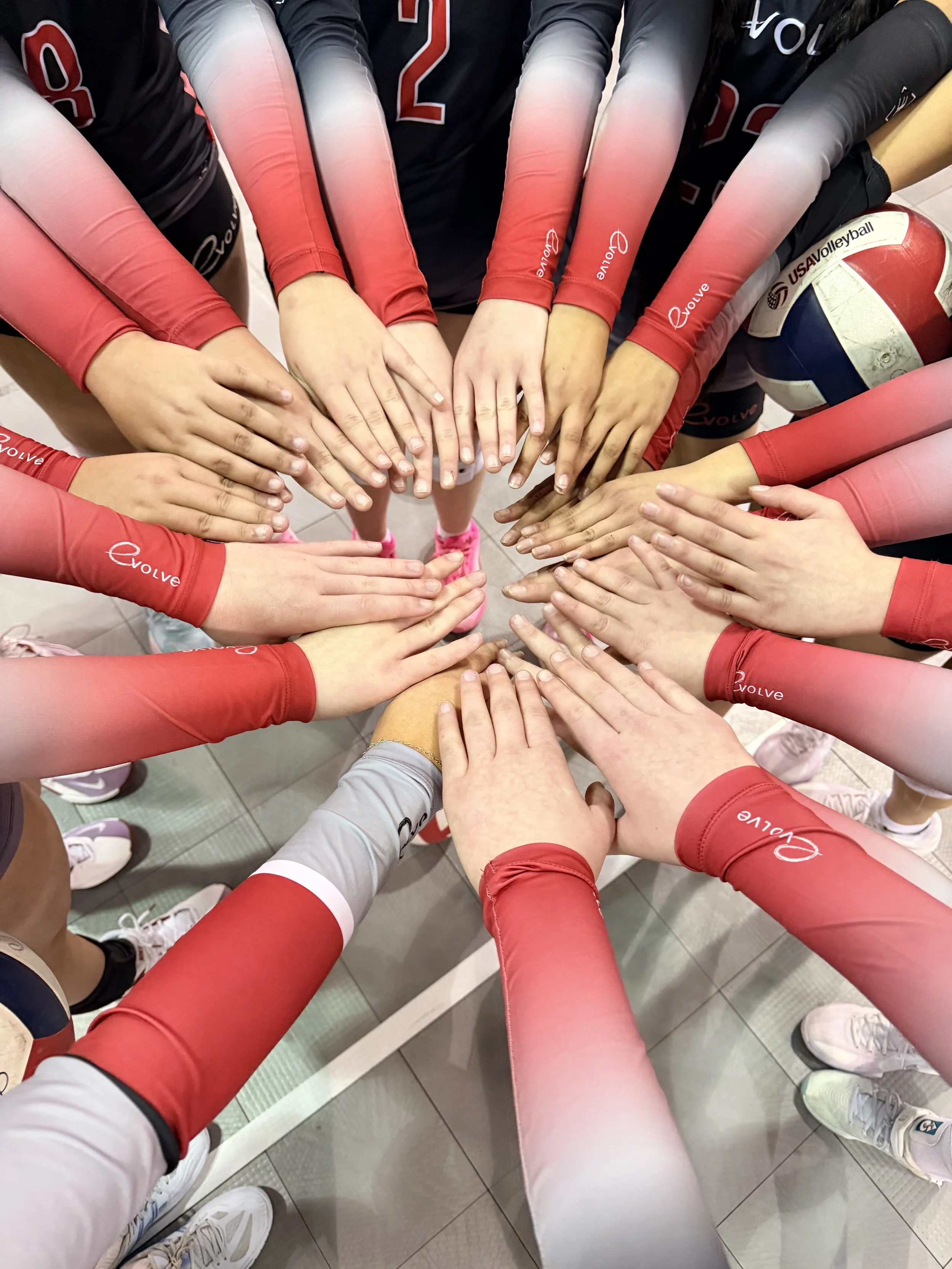 A team of volleyball players in black and red uniforms, huddled together with their hands stacked in the center, celebrating or preparing for a game, with a volleyball nearby.