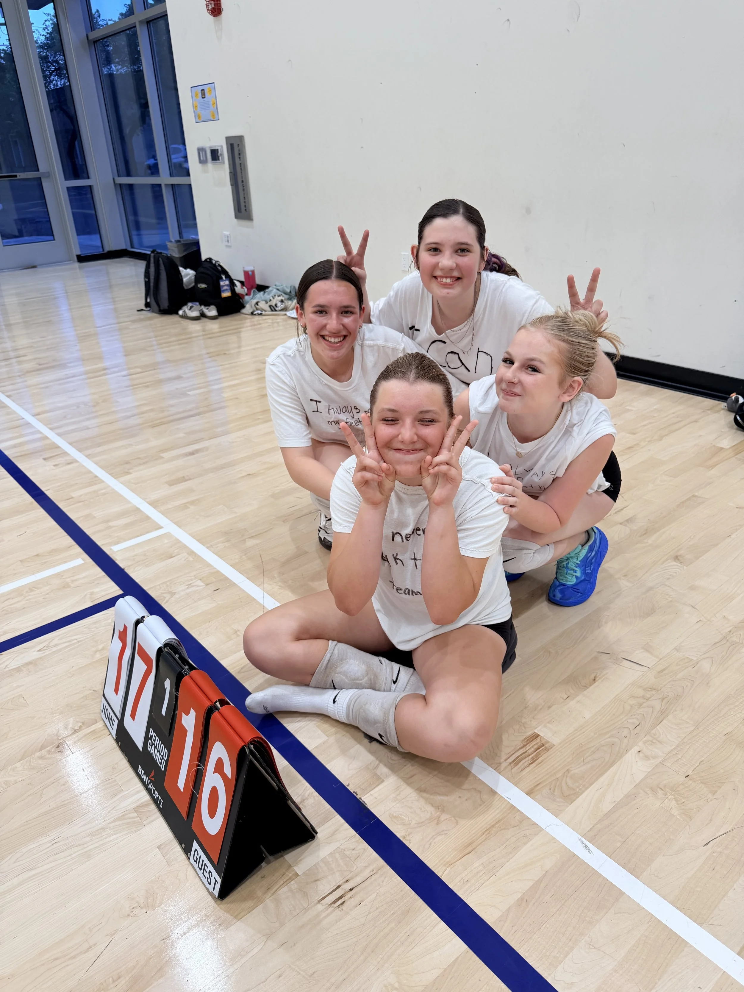 Four young girls smiling and making peace signs while sitting in a gymnasium near a scoreboard, with backpacks and shoes in the background.