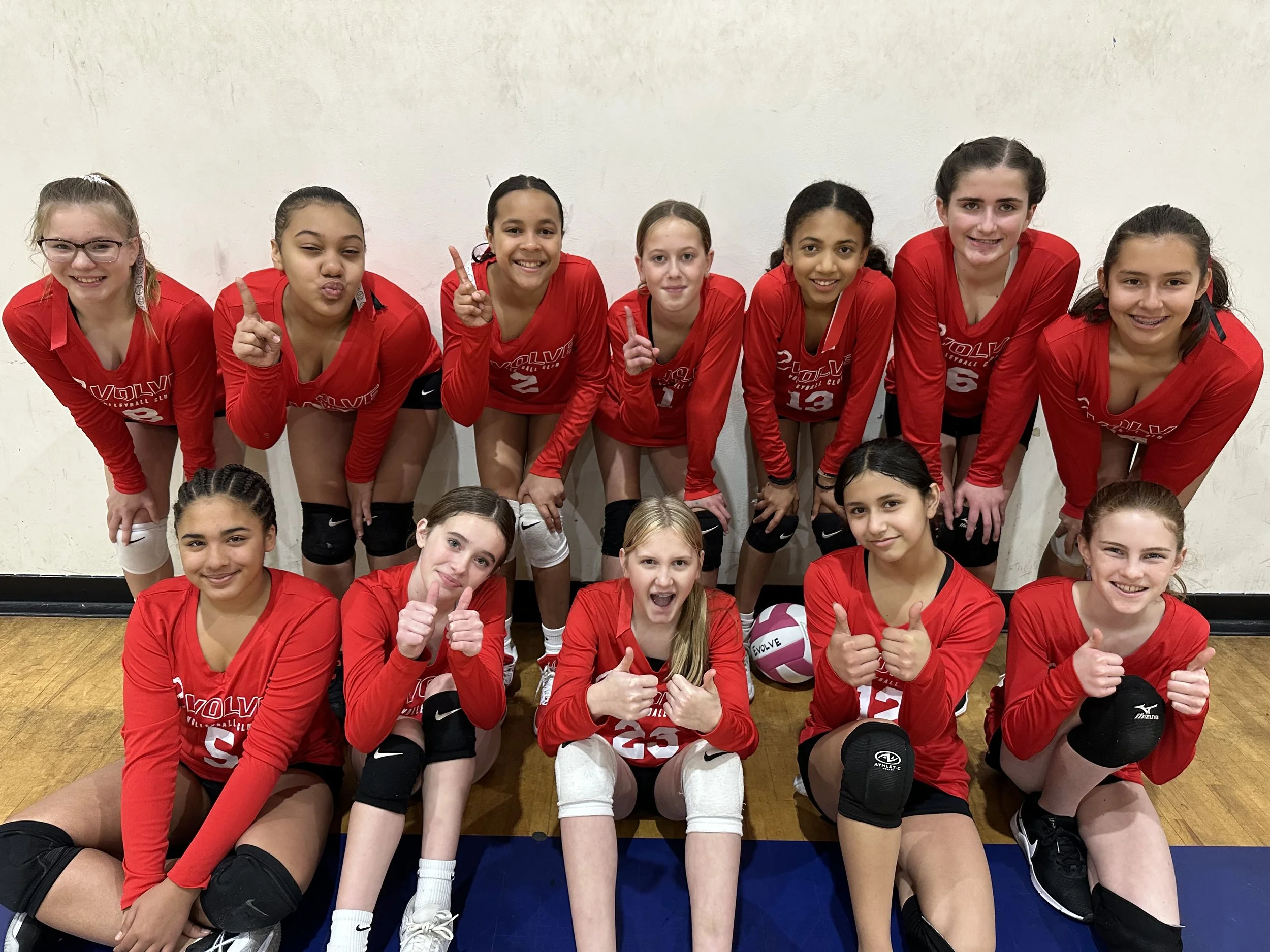 Group of young female volleyball players in red jerseys posing together indoors, some with their fingers raised or thumbs up, in front of a white wall.