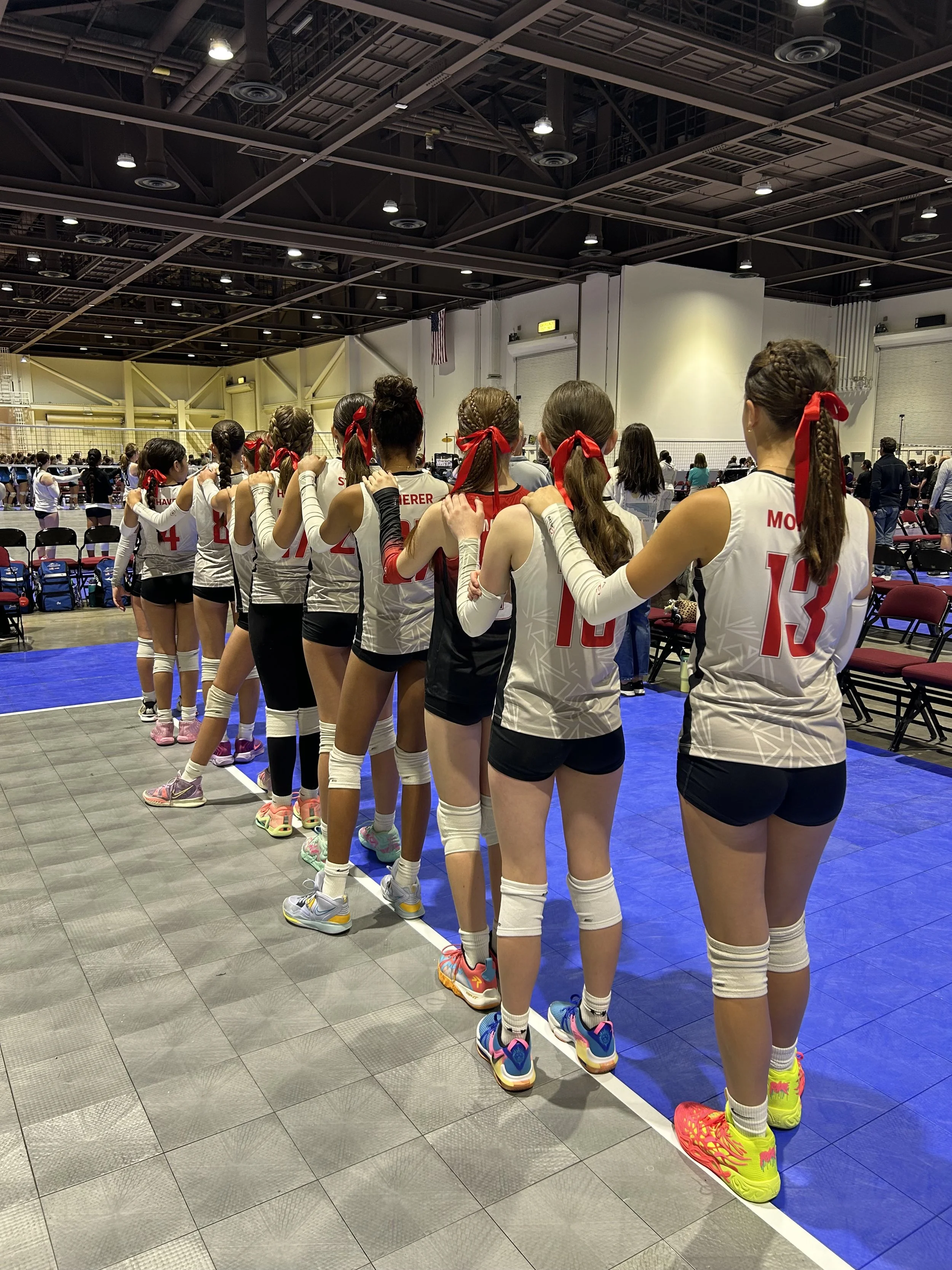 A group of female volleyball players in white and red uniforms standing in a line inside an indoor sports arena, holding each other's shoulders, with a volleyball court and spectators in the background.