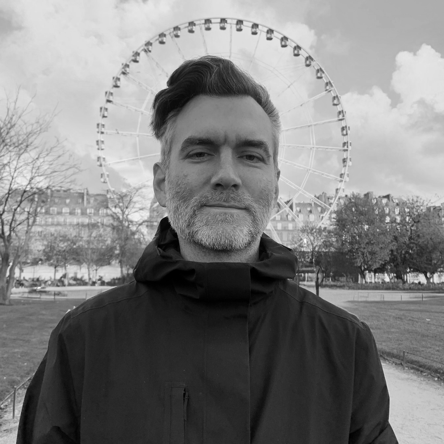 A grayscale photo of a man with a beard and styled hair, wearing a jacket, standing outdoors in front of a Ferris wheel and trees.