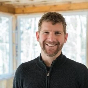 A smiling man with curly hair and a beard, wearing a black sweater, standing indoors with large windows and wooden beams in the background.