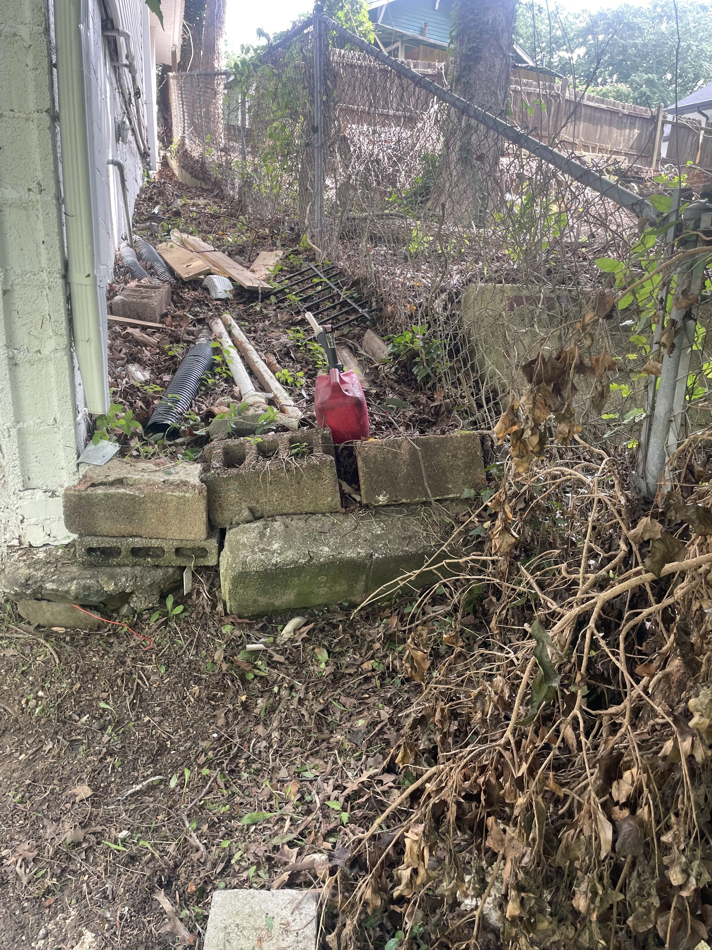A backyard with a chain-link fence, overgrown plants, a red gas can, some pipes, cinder blocks, and a concrete slab, with a house and trees in the background.