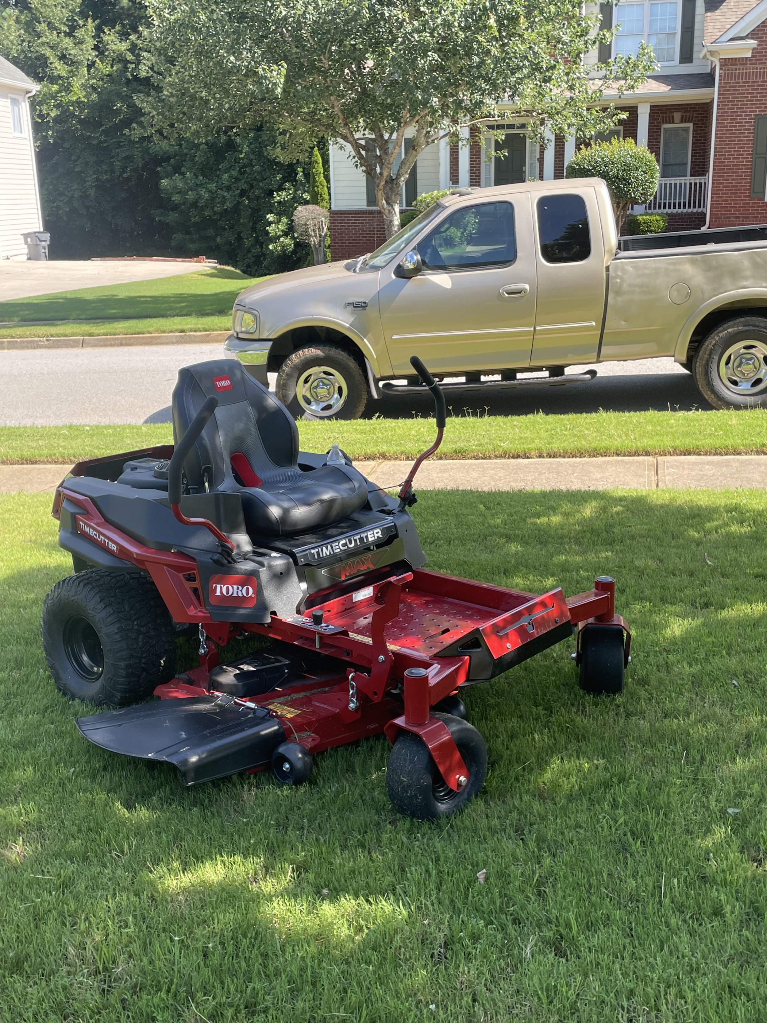 A red riding lawn mower on a grass lawn in the foreground, a silver pickup truck parked on the street, with a house, trees, and bushes in the background.