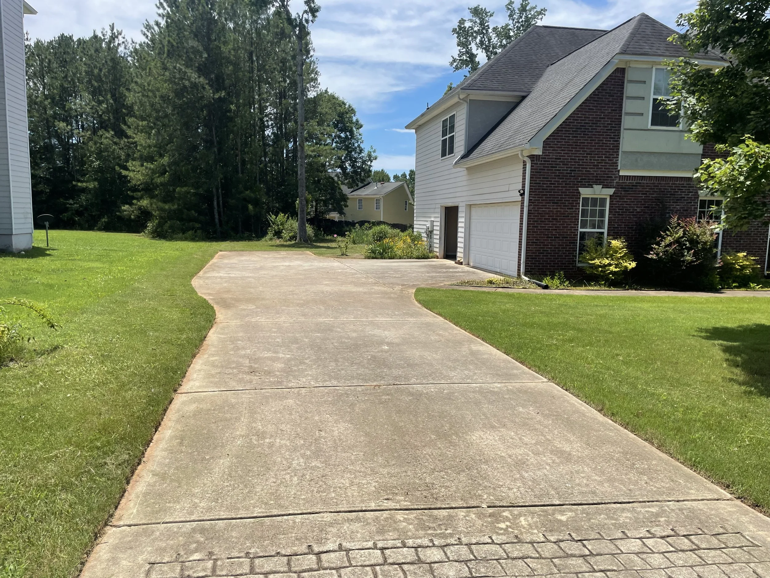 Concrete driveway leading to a garage attached to a house with white siding and brick exterior. Grass lawn on sides and trees in the background under a partly cloudy sky.