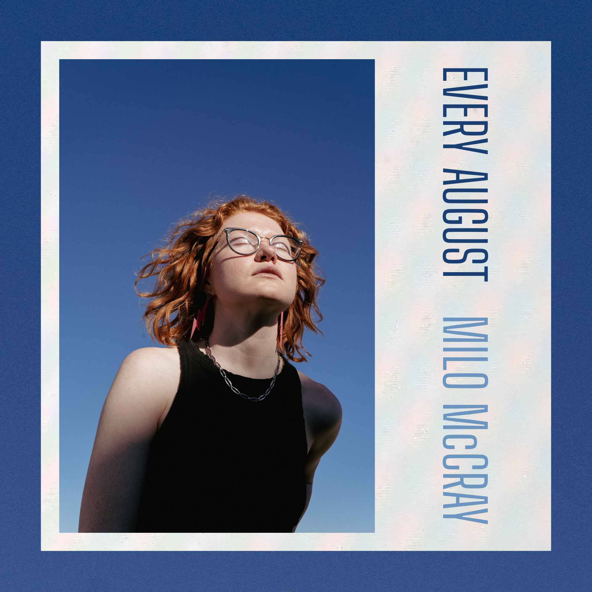 Close-up of a person with red curly hair wearing glasses and a black sleeveless top, smiling with their eyes closed against a clear blue sky.