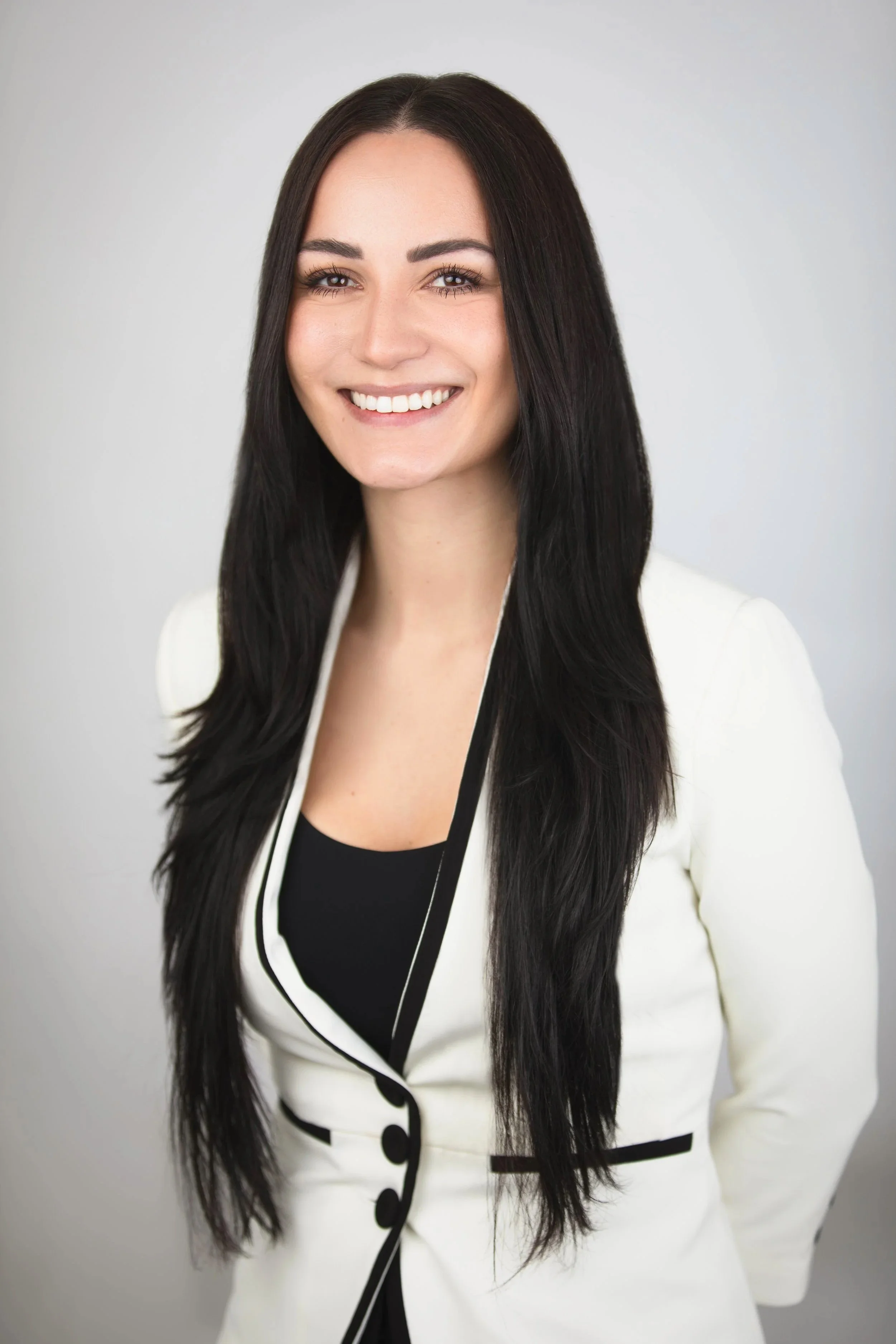 A woman with long, dark hair and a wide smile, wearing a black top and a white blazer with black trim, standing against a plain light gray background.