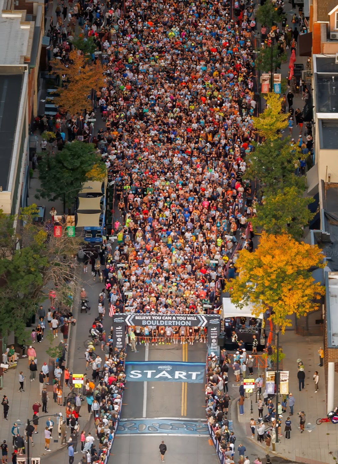 Overhead view of a large crowd gathered at the start line of the Boulderthon race in Boulder, Colorado, with colorful autumn trees lining the street.
