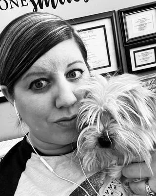 A woman taking a selfie with her dog, both looking at the camera. The woman has earphones in and is indoors, with framed certificates on the wall in the background.