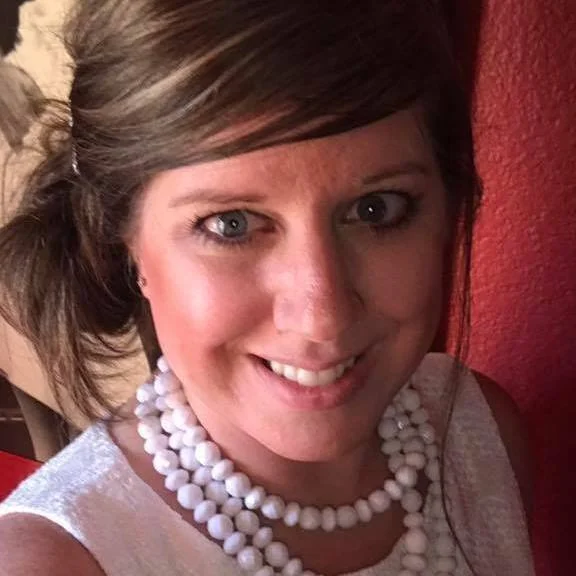 Smiling woman with light skin, blue eyes, and brown hair styled in loose waves, wearing a beaded white necklace and a sleeveless top, standing against a red wall background.