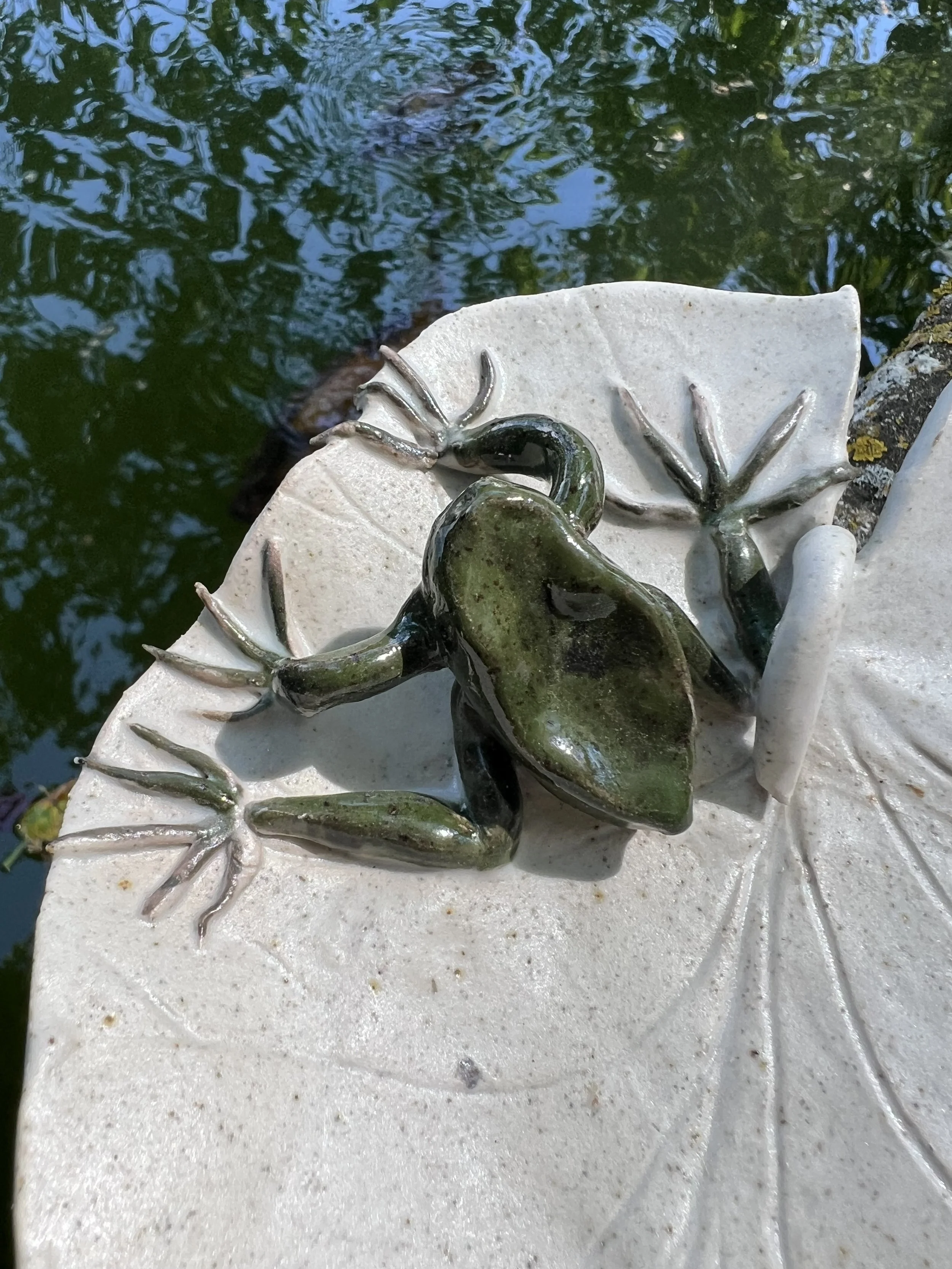 Une sculpture en céramique représentant deux grenouilles sur une feuille de lotus, posée sur une surface en pierre, avec un plan d'eau en arrière-plan.