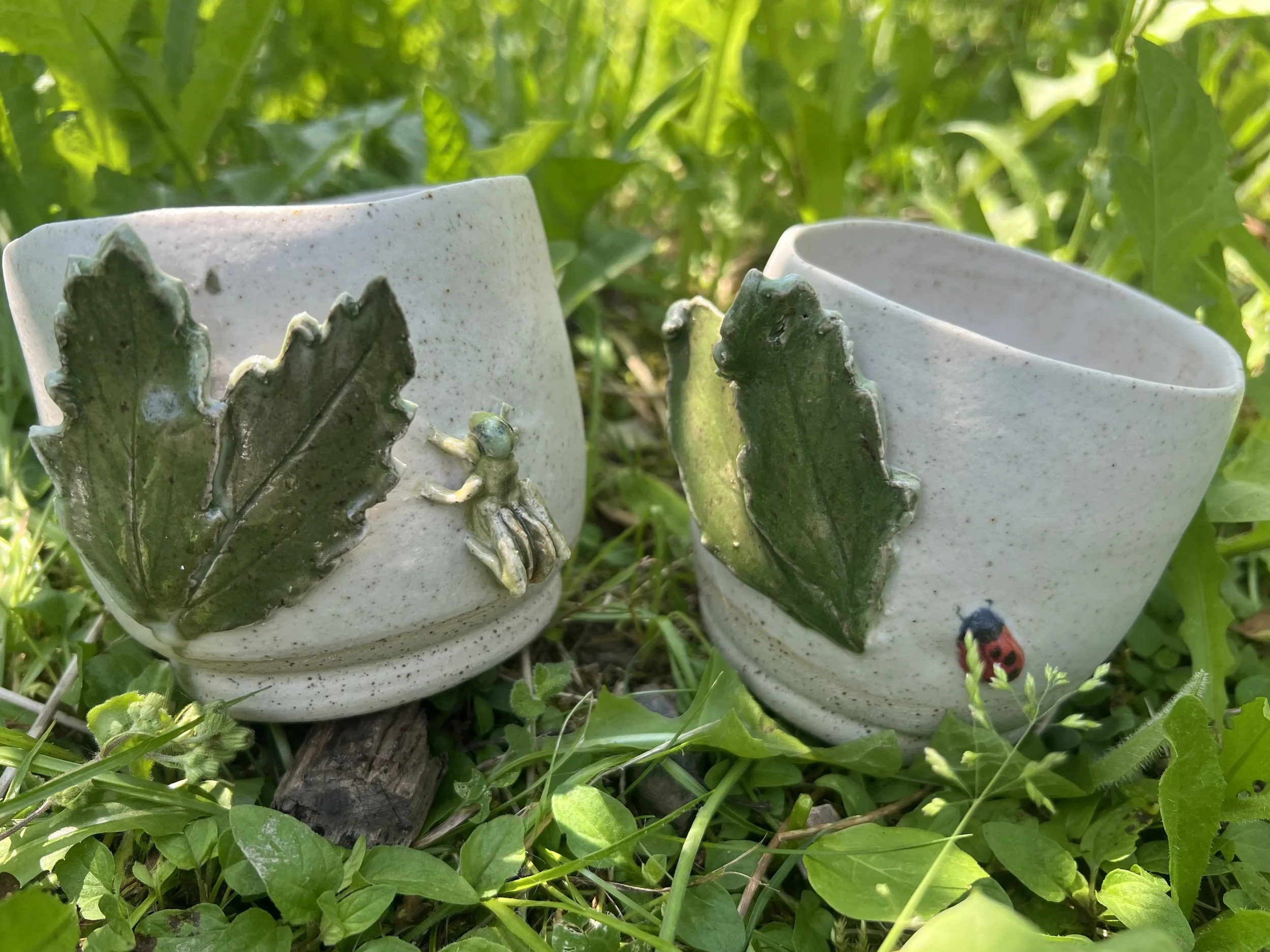 Deux mugs en céramique avec des décorations en forme de feuilles vertes sont placés dans l'herbe, entourés de végétation dense. Un petit insecte vert est posé sur l'une des feuilles et une coccinelle rouge est visible au bas à droite.