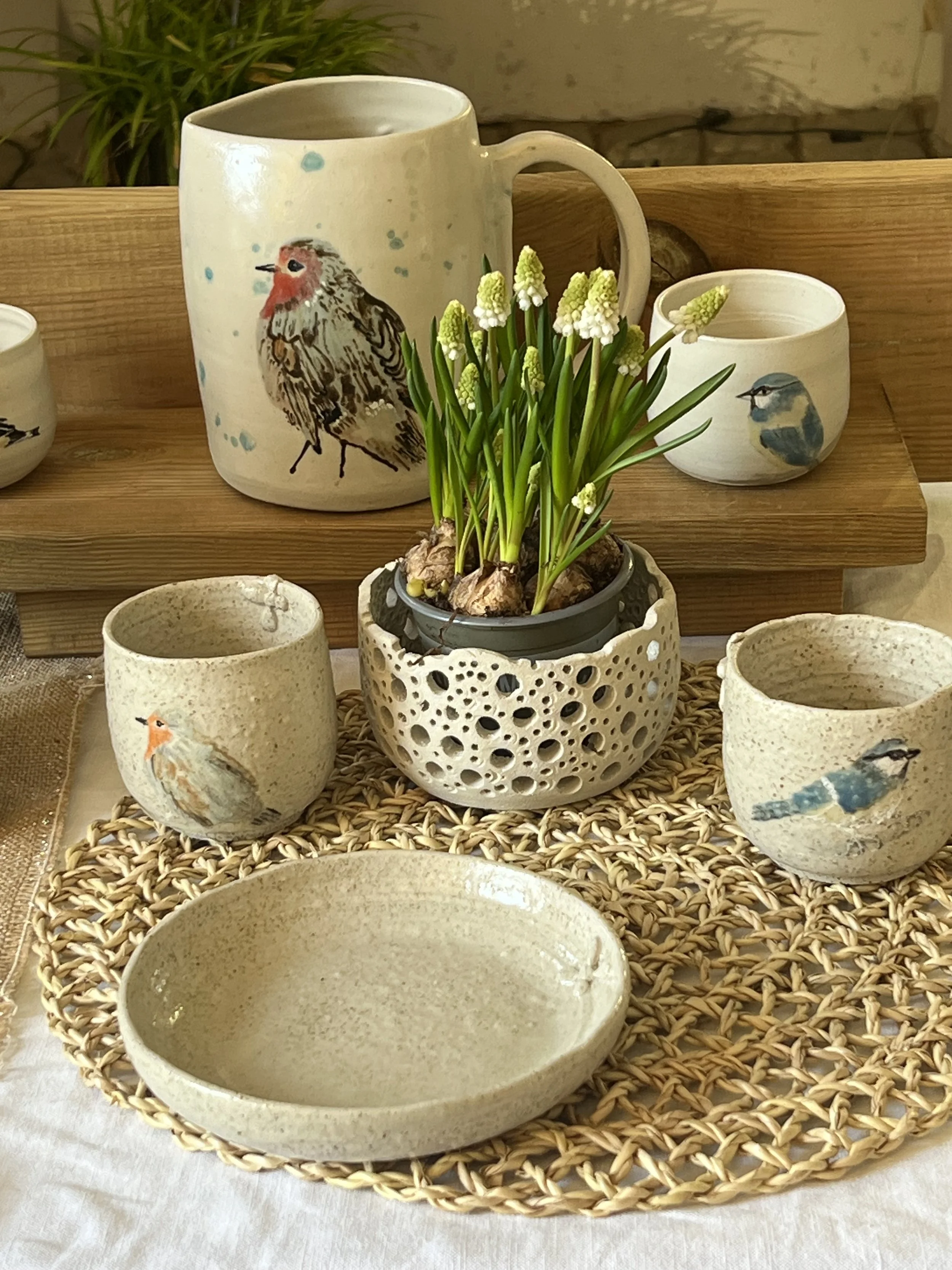 Une collection de porcelaine comprenant un mug, plusieurs tasses et plats, décorés de peintures de petits oiseaux, tous placés sur un plateau en rotin, avec une plante verte en arrière-plan.