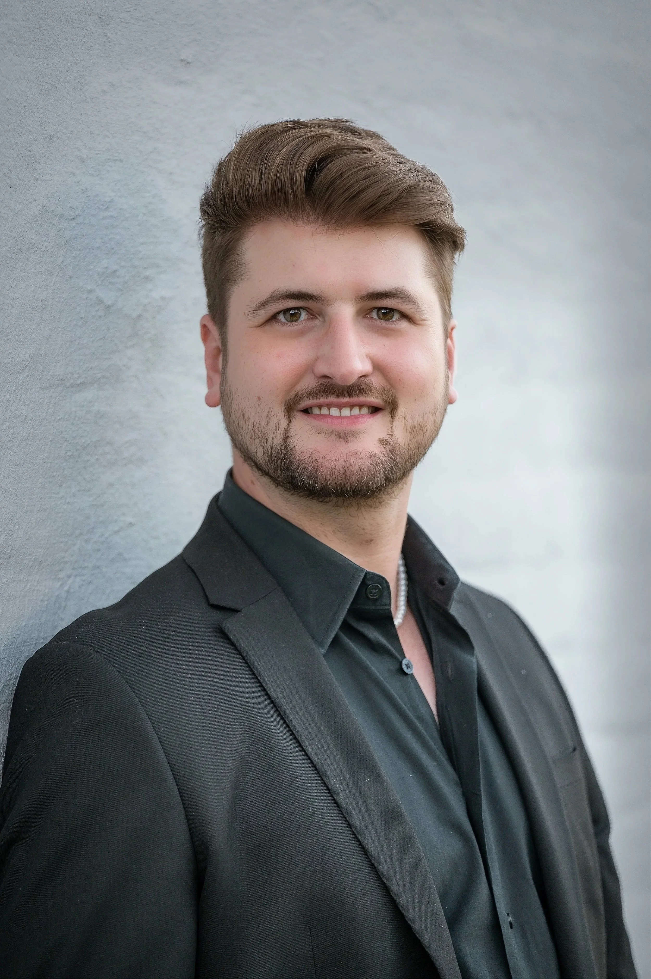 A young man with light brown hair and a beard, wearing a black suit and shirt, smiling while standing against a light grey wall.