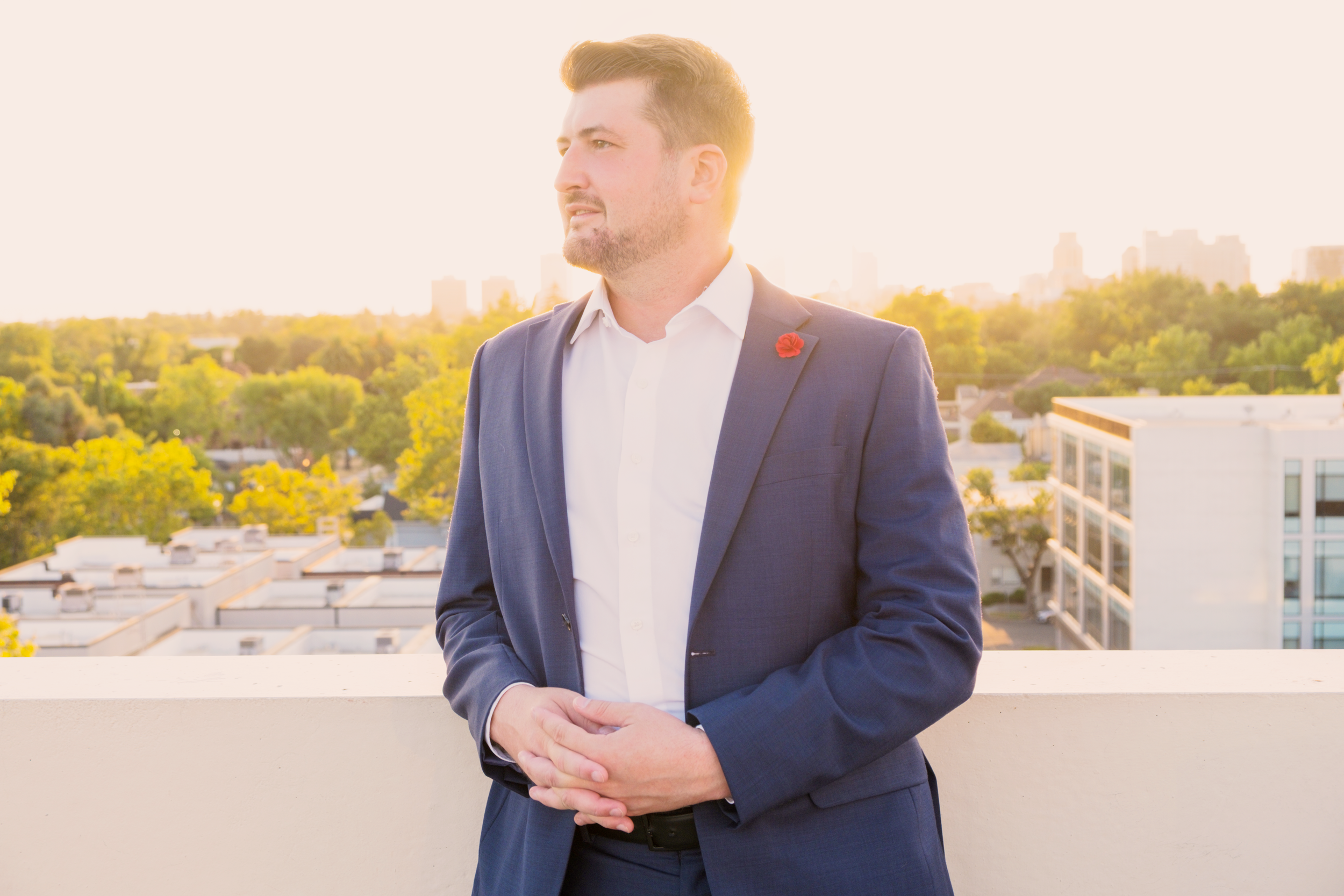 A man in a dark suit and white shirt standing on a balcony during sunset, with a cityscape and green trees in the background.