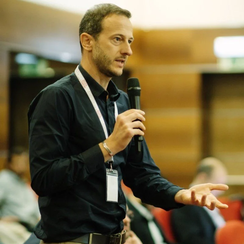 A man with short brown hair and a beard, wearing a black shirt, speaking into a microphone at an indoor event.