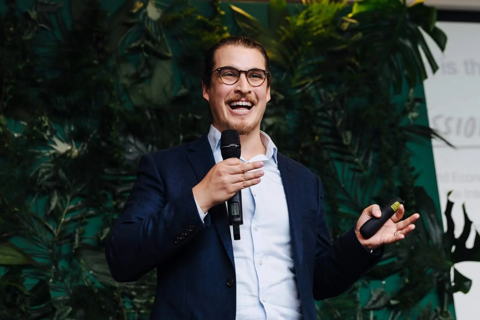 A man in a suit and glasses smiling while holding a microphone and a remote control, standing in front of green plants.