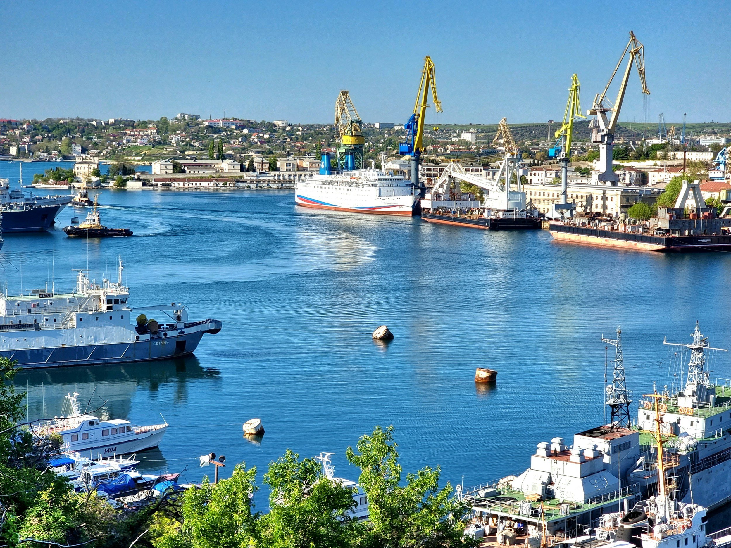 A harbor with various boats and ships docked, along with large industrial cranes, with a hillside town in the background.