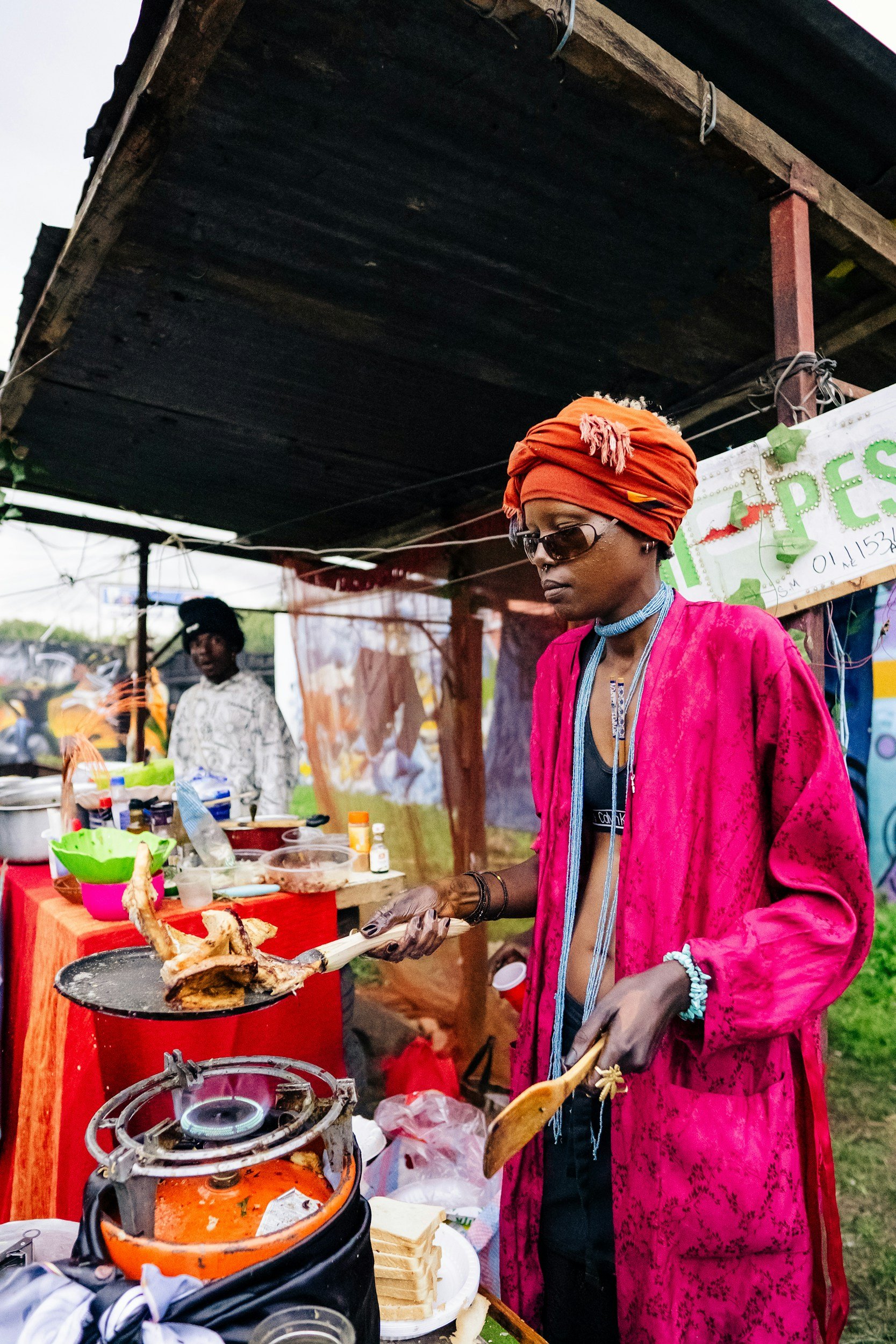 A woman wearing sunglasses, a red headscarf, and jewelry is cooking food at an outdoor stand with a red table. She is holding a spatula with a piece of food on it. There are various food items and cooking supplies on the table, and another person is visible in the background.